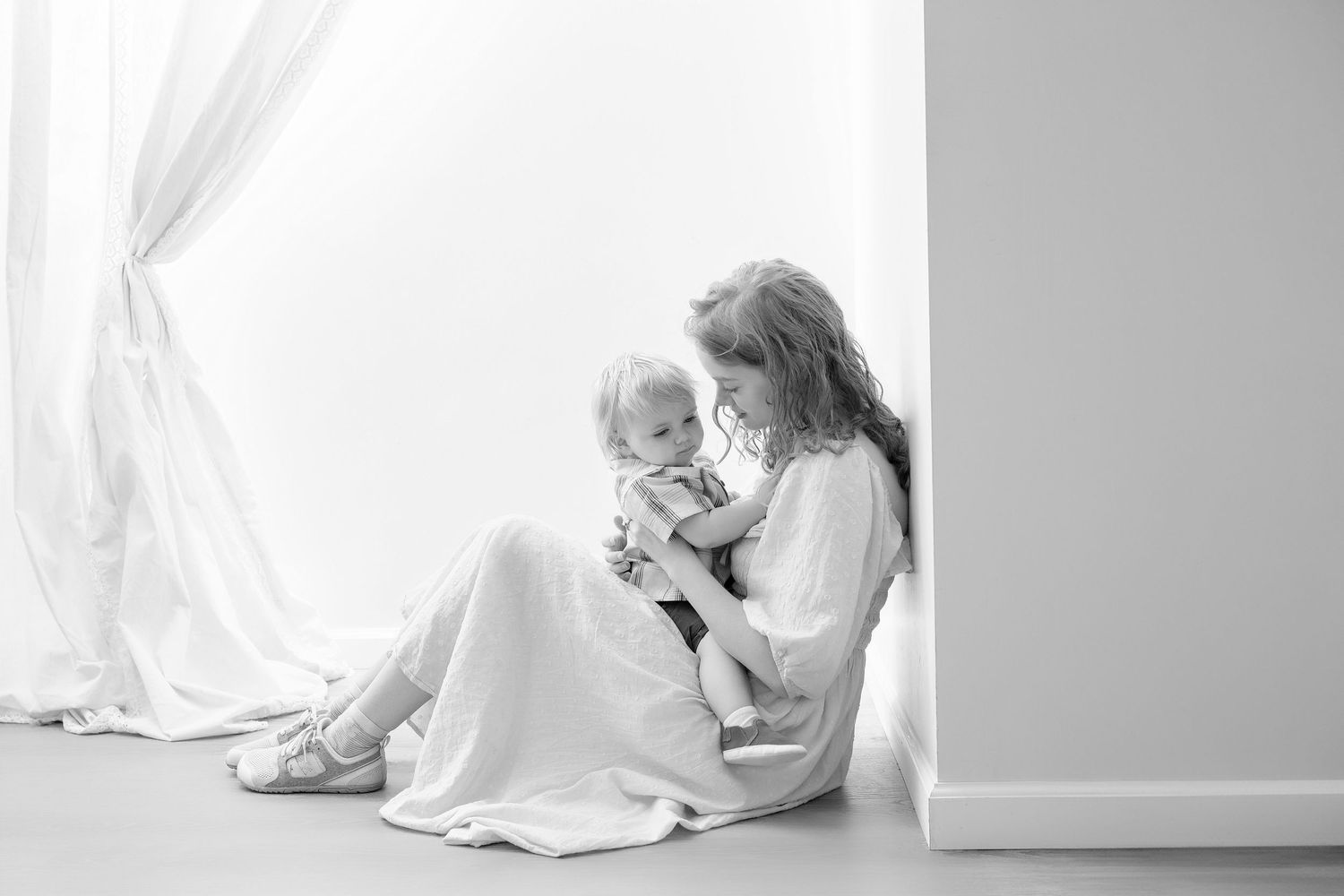 Black and white photo of a mother sitting on floor holding her baby near a bright window.. Minnesota milestone photographer