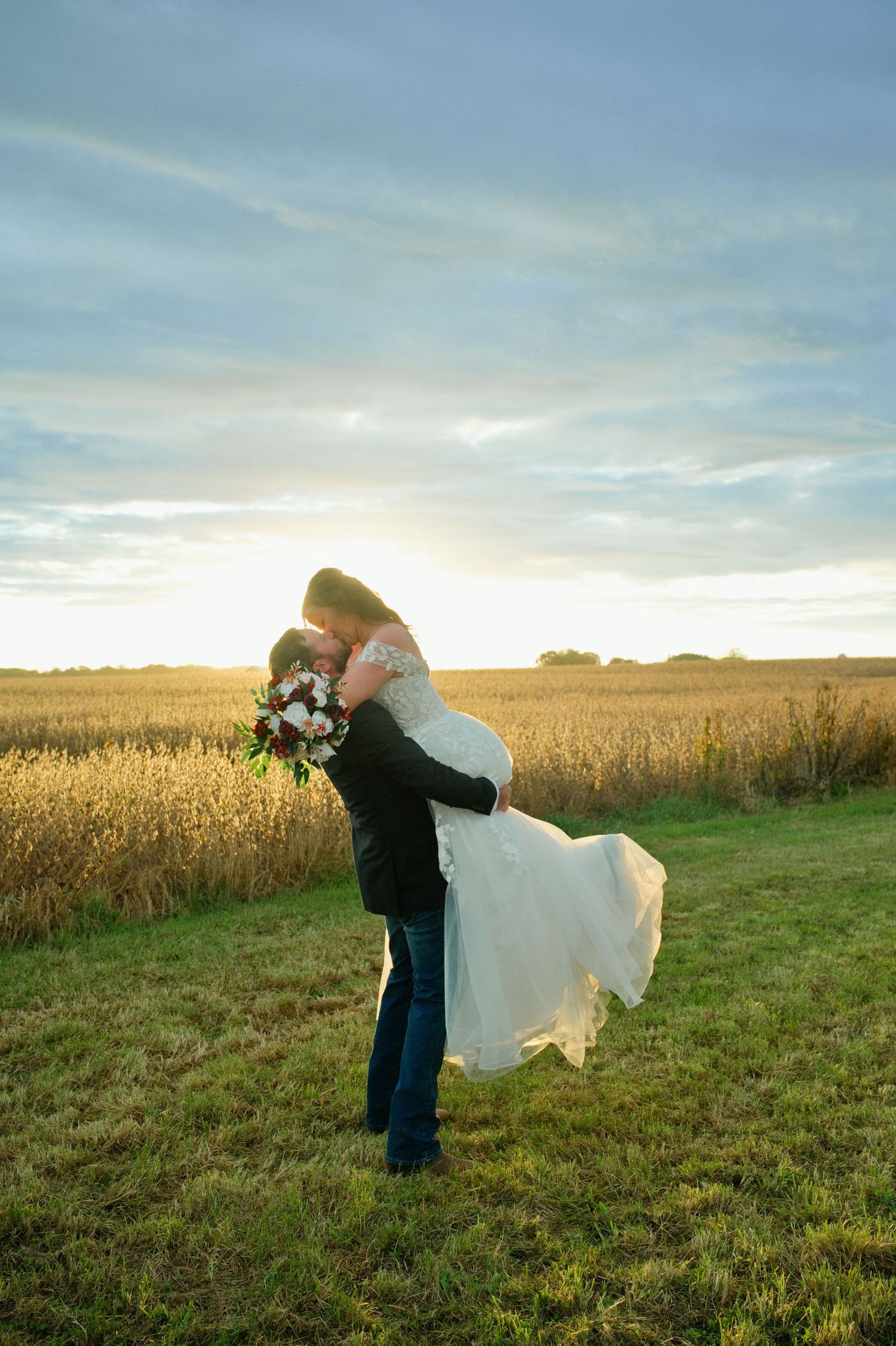Groom lifting bride in white dress at sunset in golden field, romantic outdoor wedding photo.