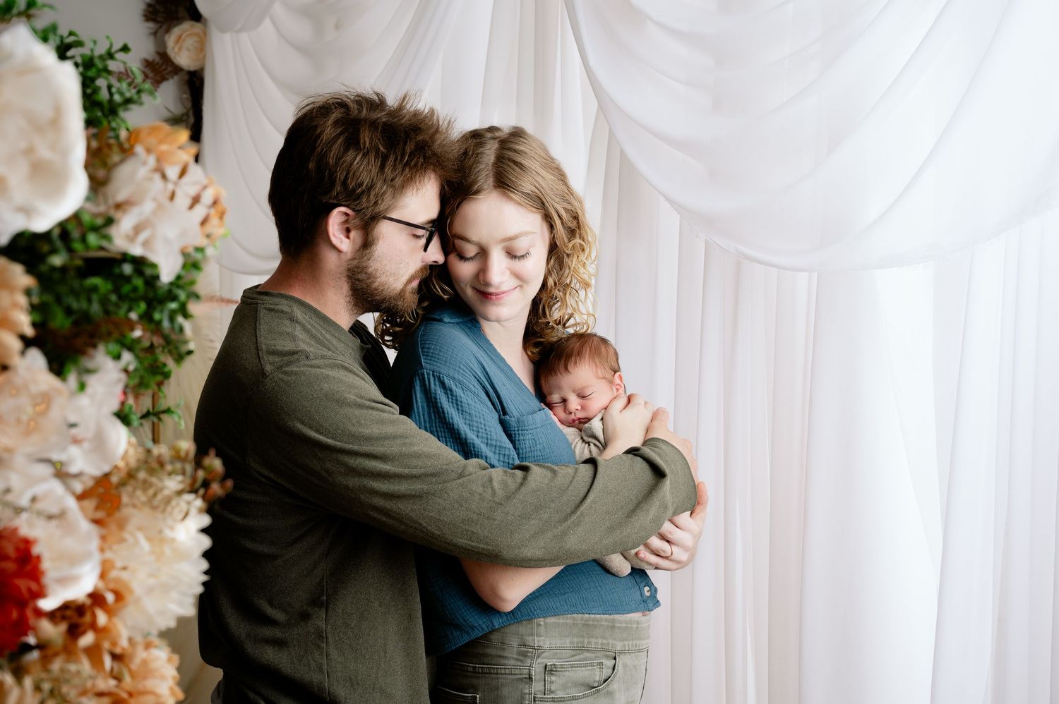 St Cloud Best newborn photographer - A romantic embracing couple with newborn in a light-filled room with a floral backdrop.