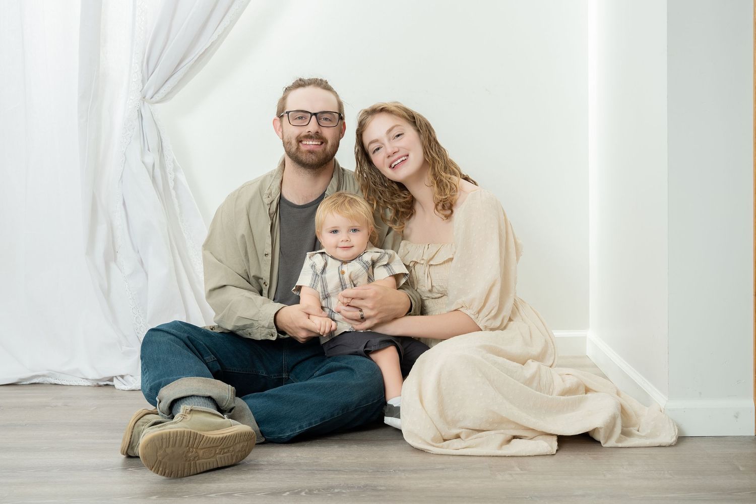 Family of three poses in matching neutral outfits during cozy indoor photo session with white backdrop. Princeton MN Milestone, Maternity an