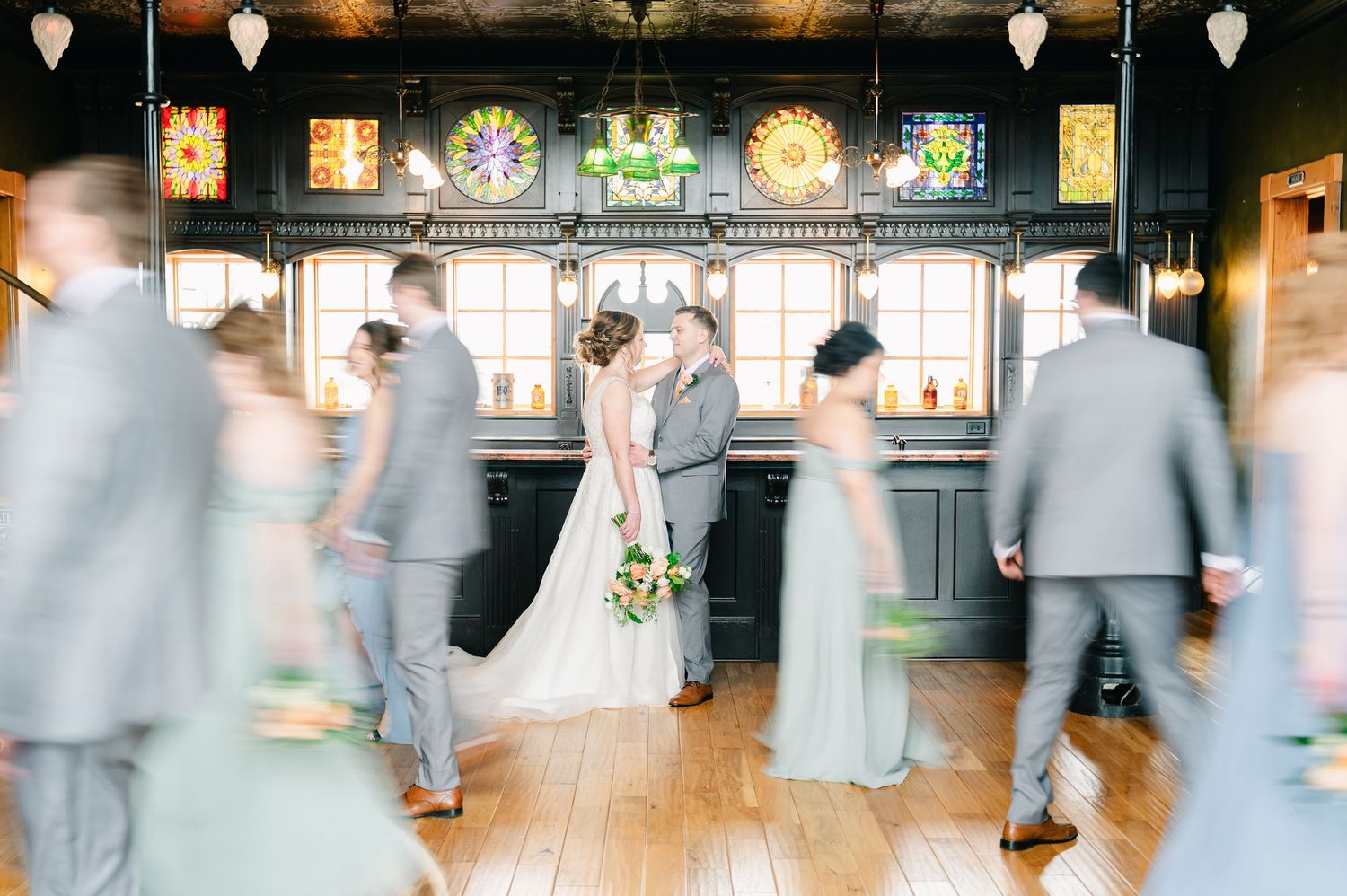 Bride and groom kiss surrounded by motion-blurred wedding party in elegant stained glass venue.