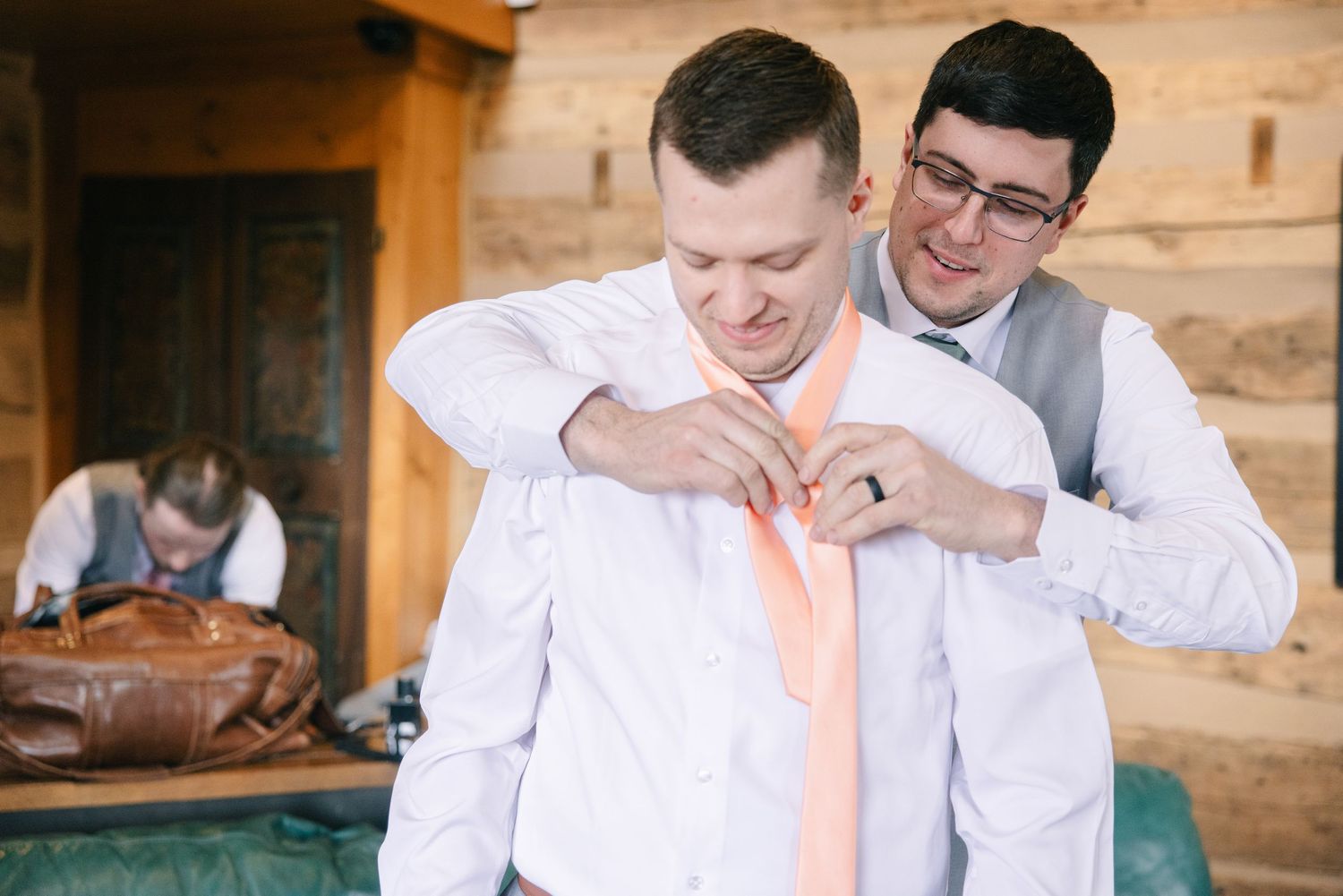 Groomsman helping groom adjust peach tie during wedding preparations in rustic wooden venue.