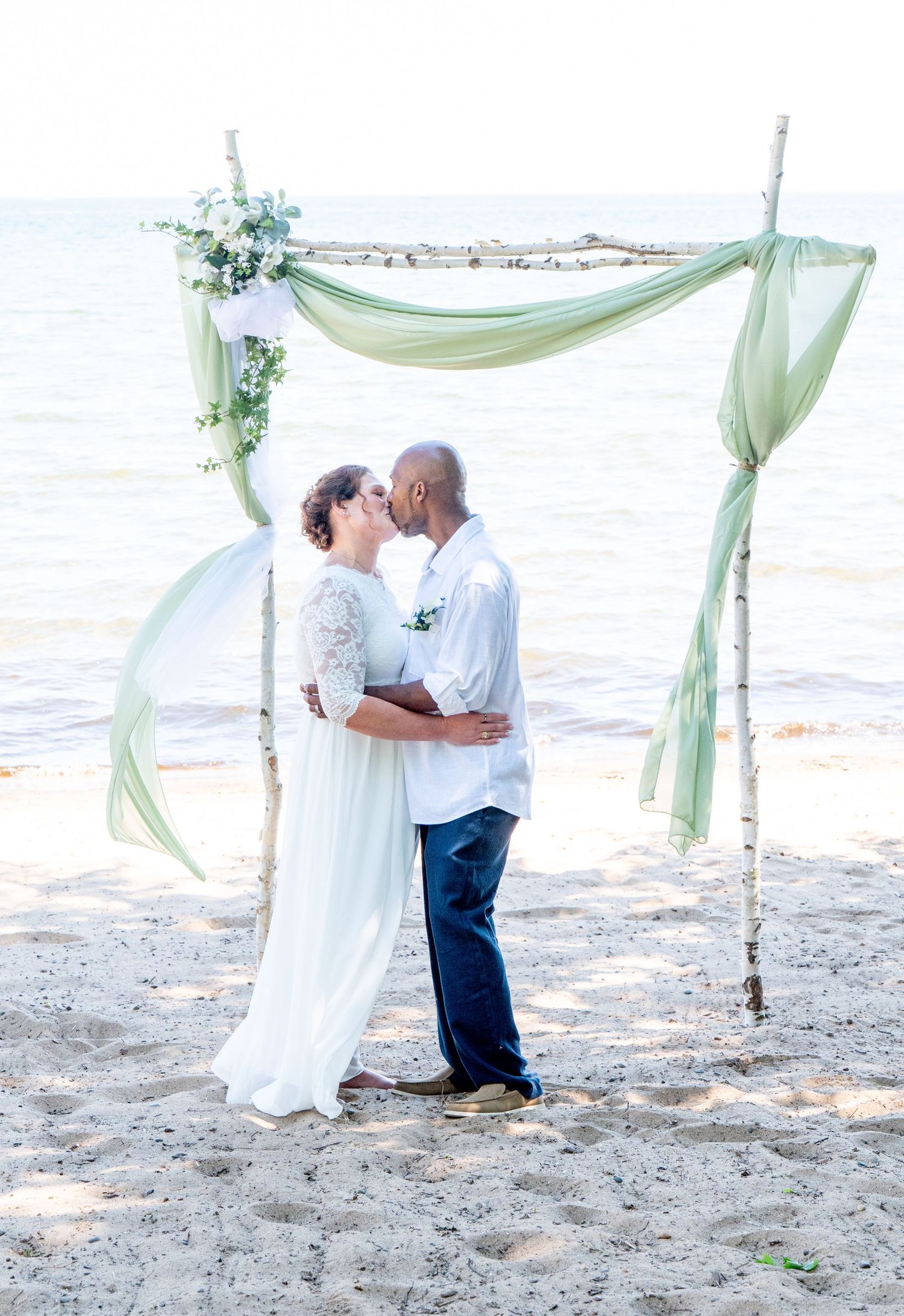 A couple shares a kiss under a driftwood arch draped in sage green fabric on a sandy beach.