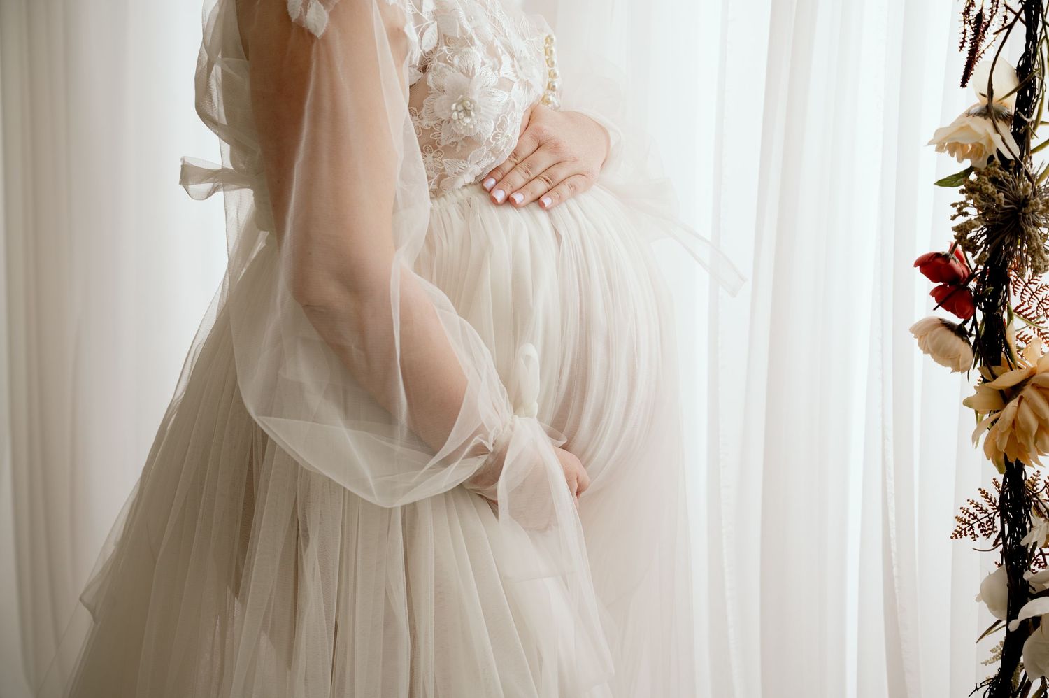 Close-up of a delicate white tulle wedding gown with lace details against sheer curtains in soft natural light.