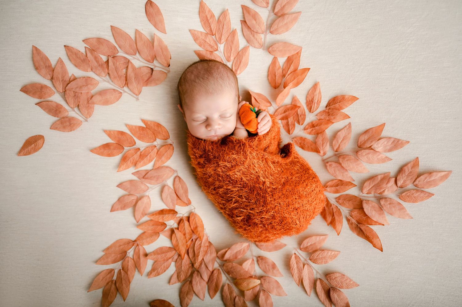 A baby lies on white fabric surrounded by orange autumn leaves arranged in a decorative circular pattern.