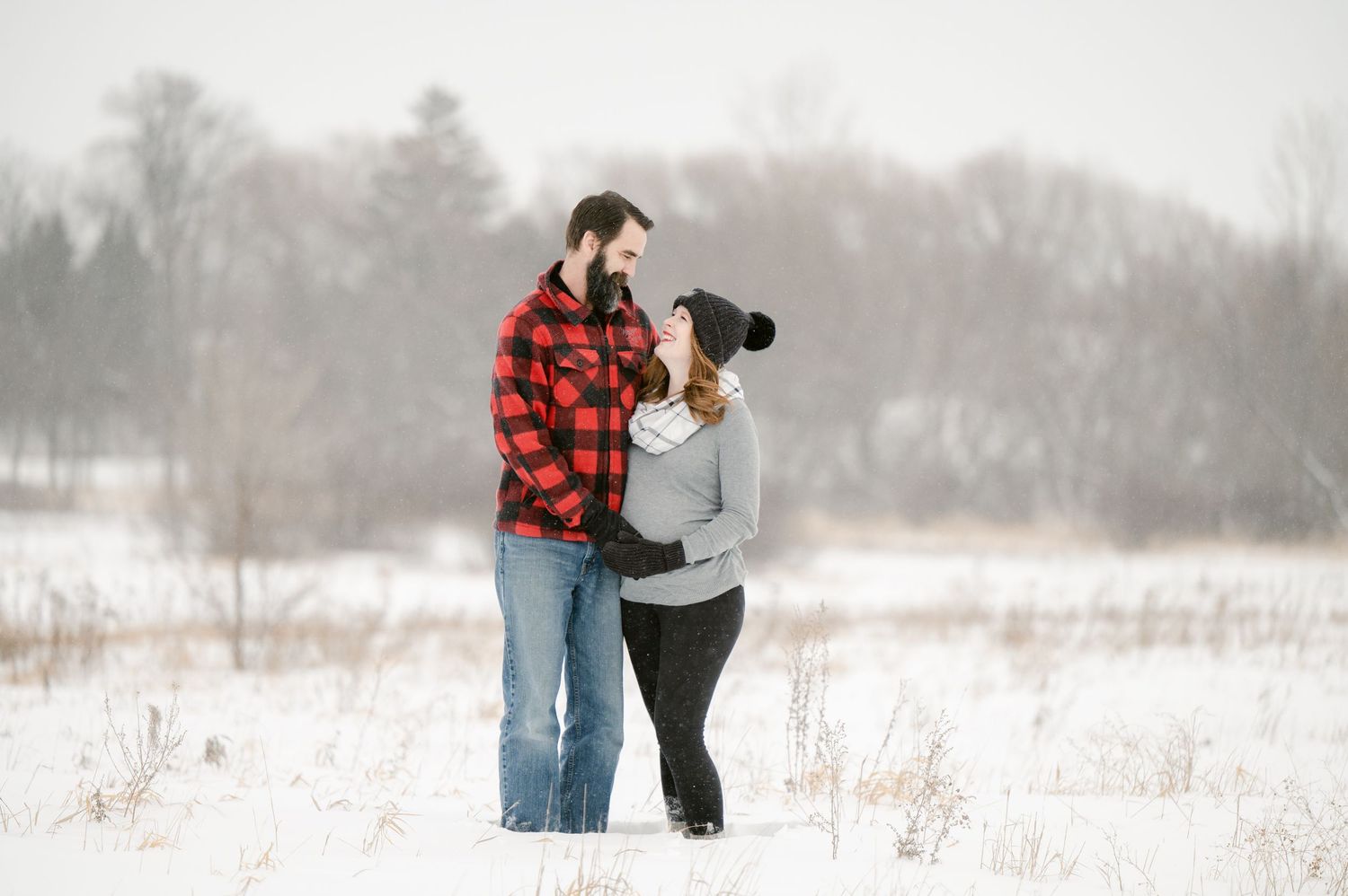 The Clark’s romantic winter maternity photoshoot series in Foreston MN, showing a couple cuddling in a snowy field wearing plaid and white.