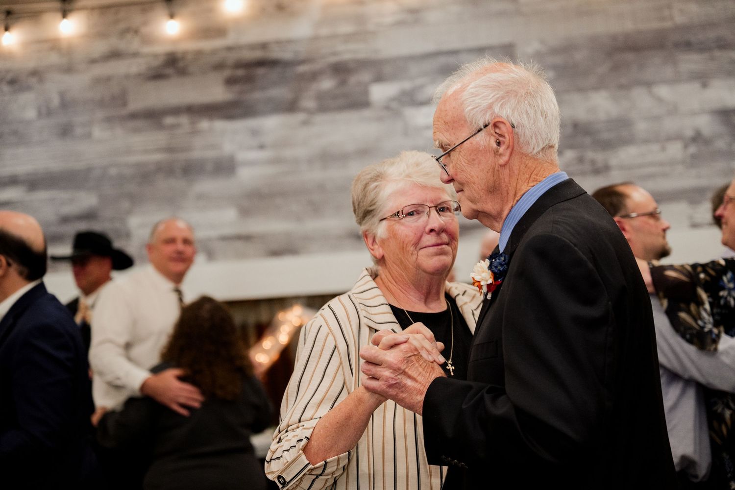 Couples dancing together at an indoor event with rustic wooden walls and string lights.