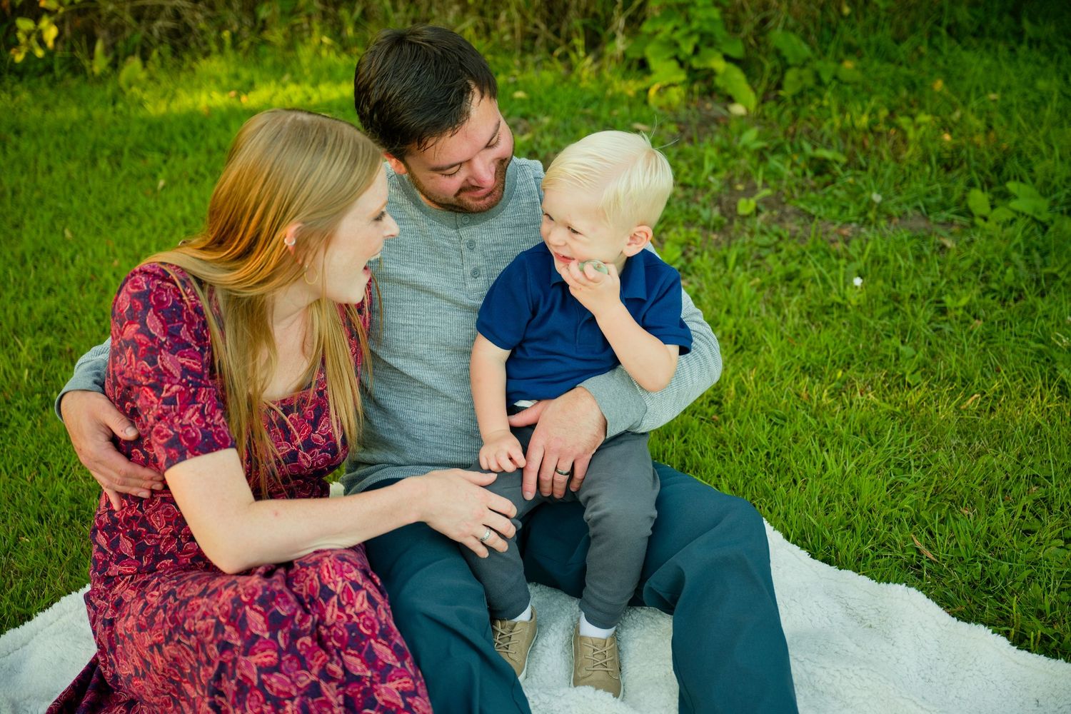 A young family sits on a white blanket in green grass, parents smiling at their blonde toddler son.