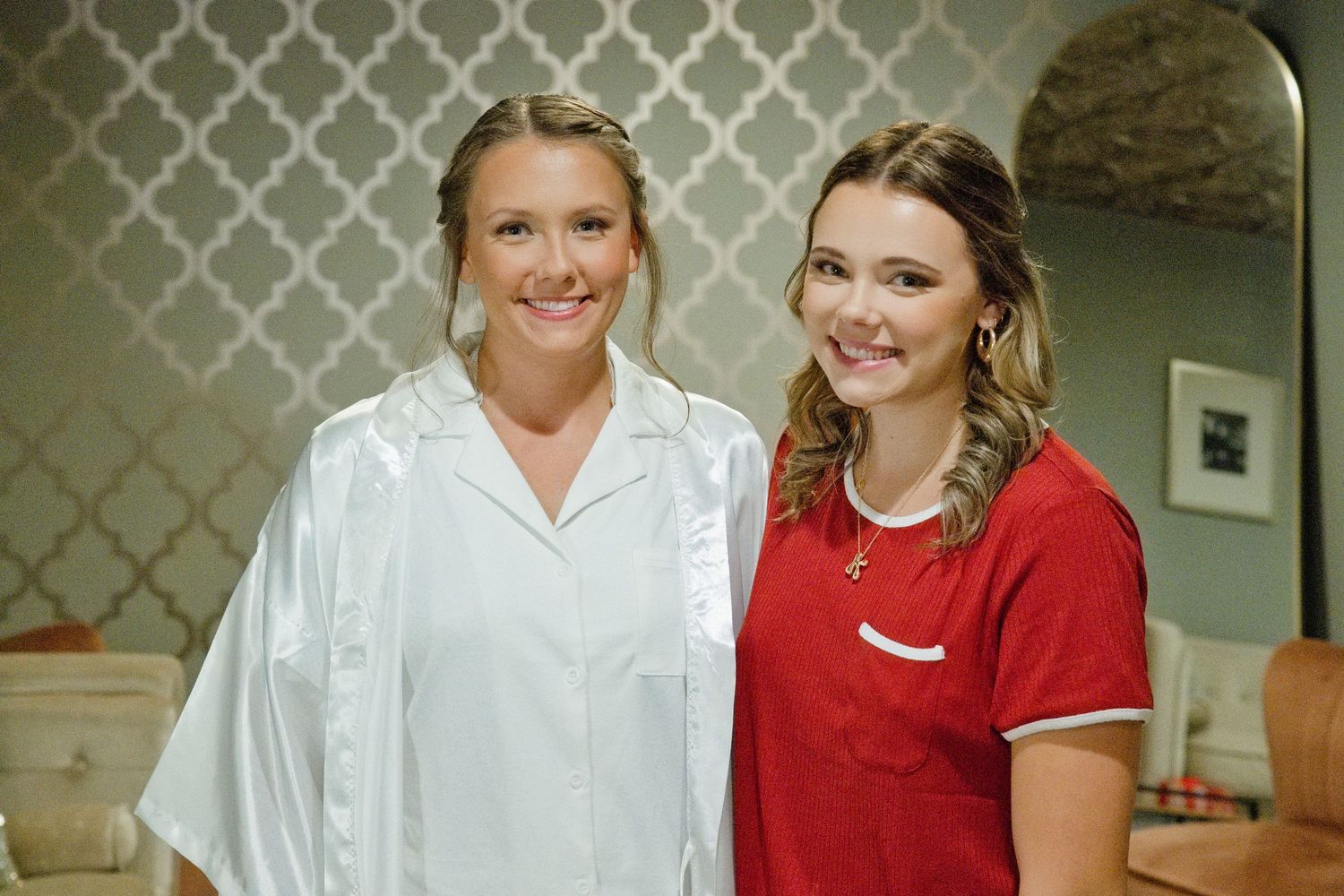 Two smiling young women in a bridal suite, one in a white satin robe and one in red.