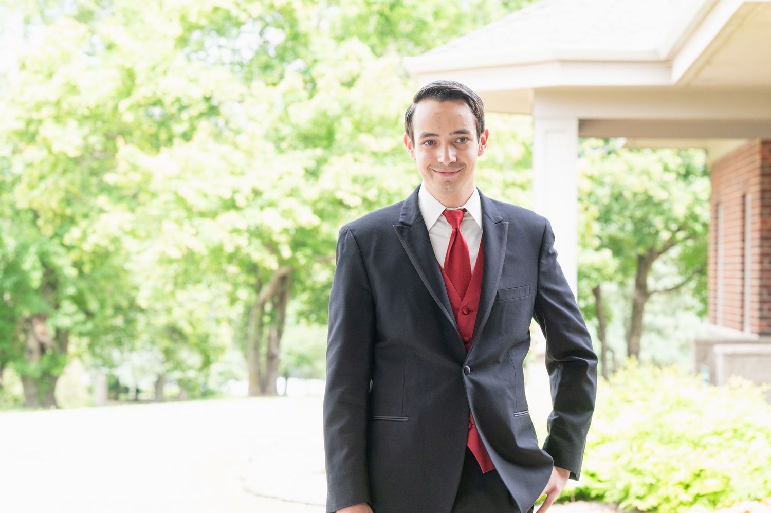 Handsome groom in dark suit and red tie smiling outdoors among lush green trees on wedding day.
