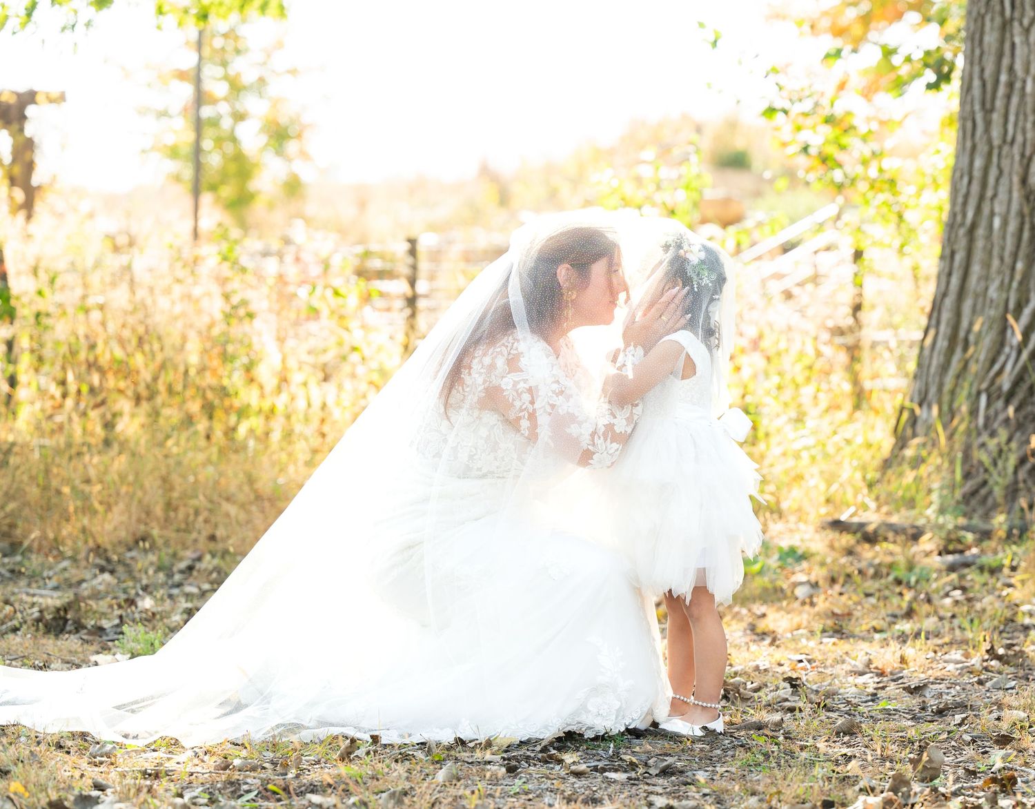 Bride in white lace gown and veil kneels to embrace a young child outdoors in autumn foliage.