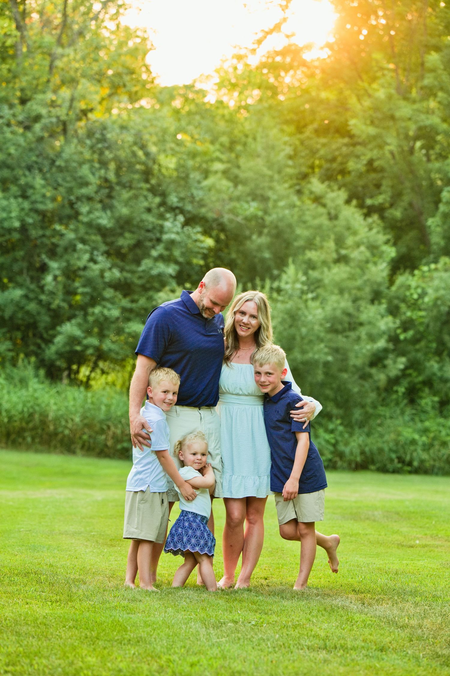 Family of five posing outdoors on green grass with sunlit trees, parents and three young children smiling together.