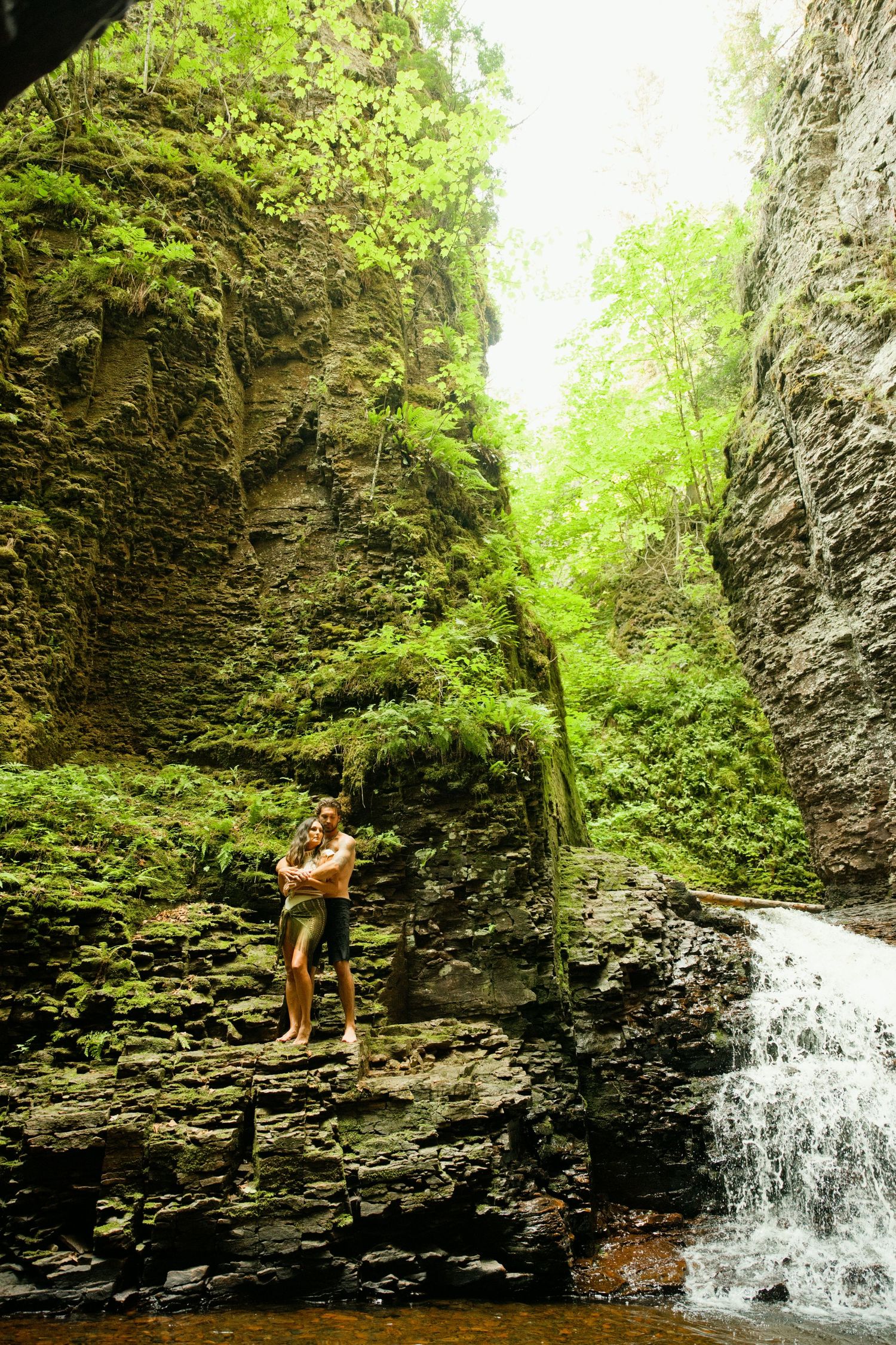 Couple embracing on rocky ledge beside a cascading waterfall in a lush green canyon.