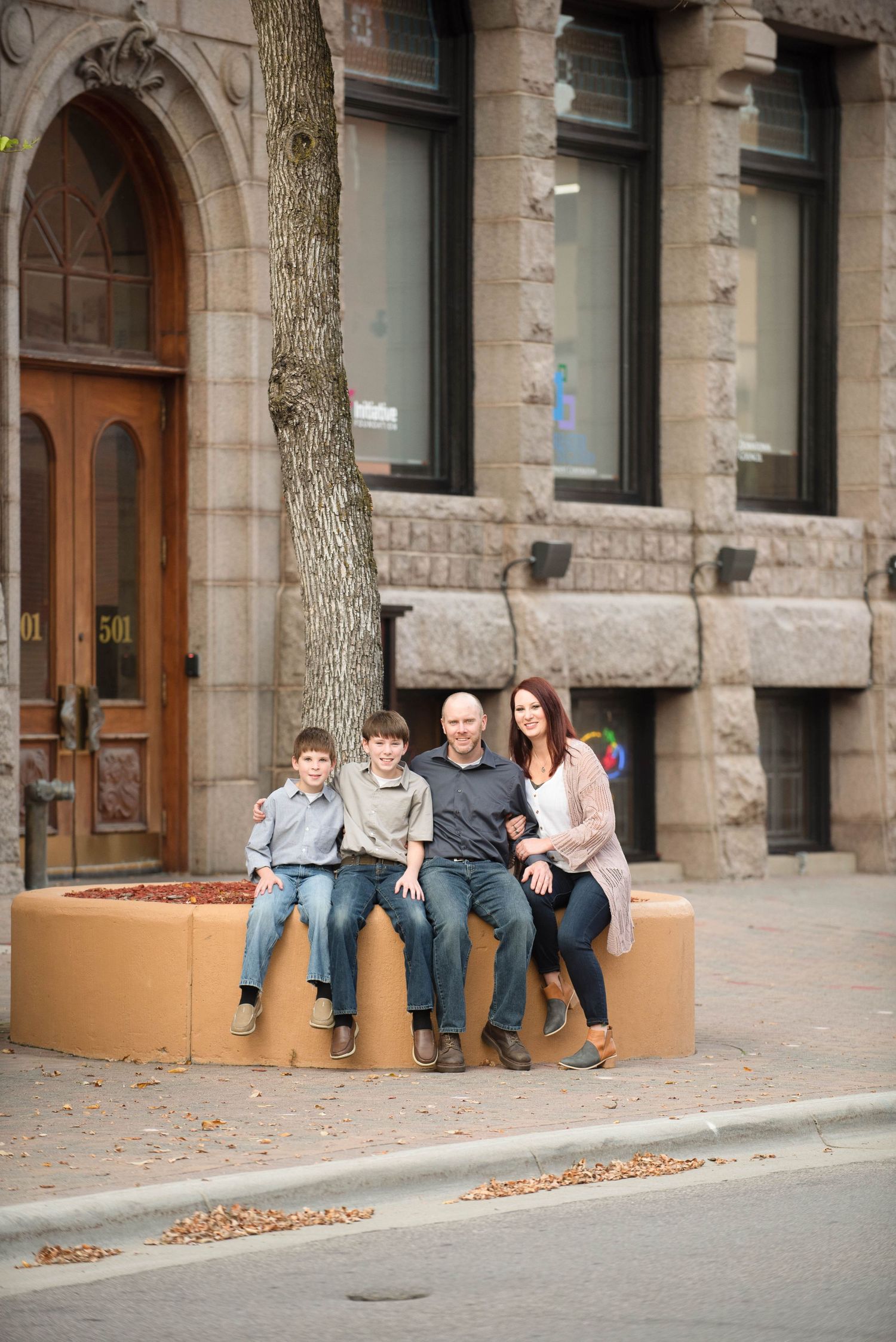 People sit casually along a stone building's exterior planters on an urban sidewalk with fallen autumn leaves.