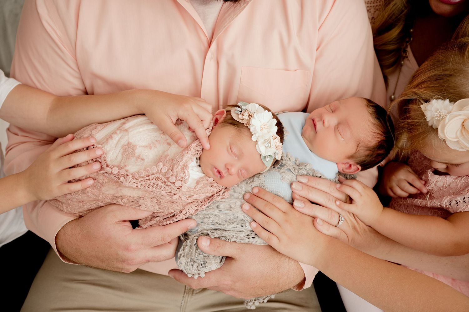 Parents tenderly hold sleeping twin newborns wrapped in lace, with an older sibling nearby in a family portrait.