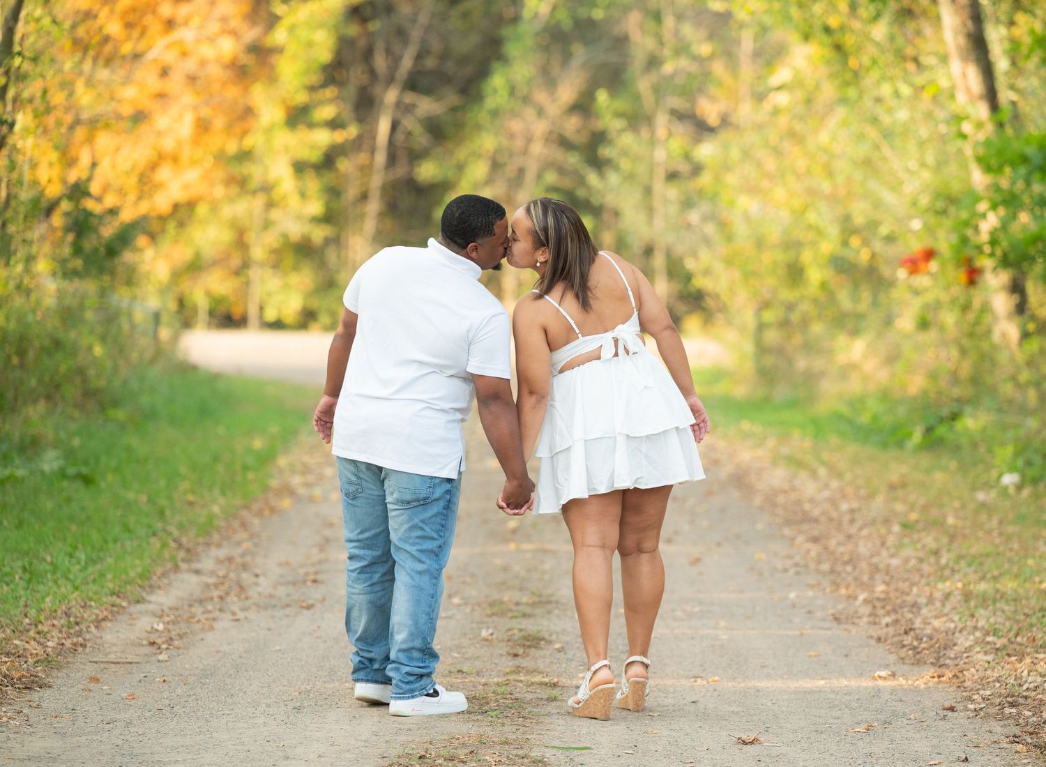 Couple kissing on a tree-lined path in autumn, holding hands, woman in white dress, man in white shirt and jeans.