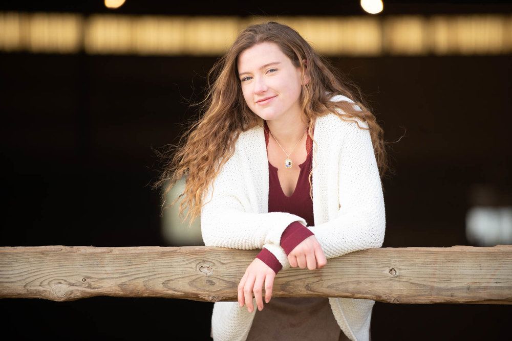Central MN Senior Photography - Young woman with curly hair smiling, leaning on a wooden fence rail outdoors in a white cardigan.