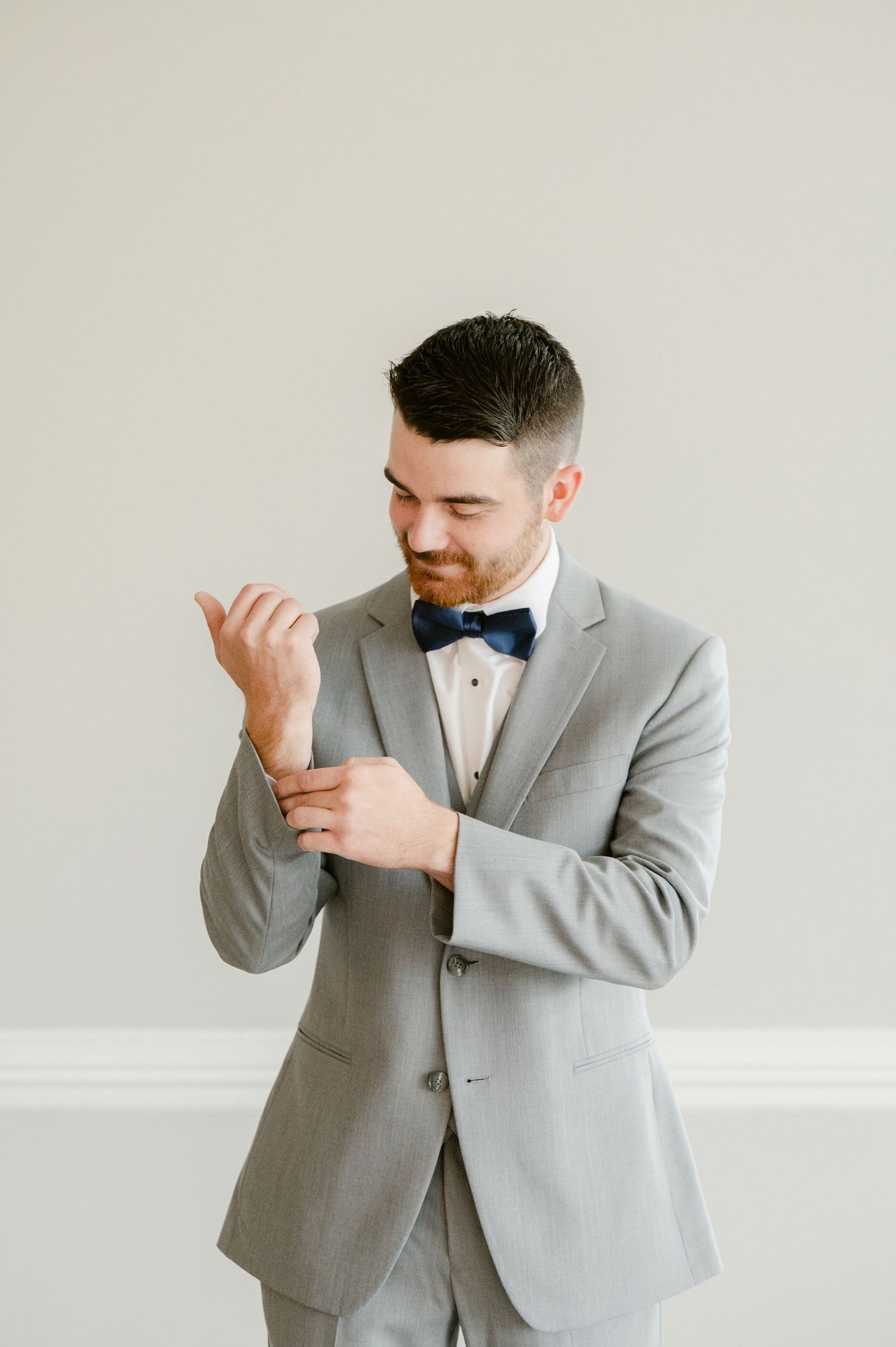 Central MN Wedding Photos | Groom in gray suit and navy bow tie adjusting cufflinks while smiling.