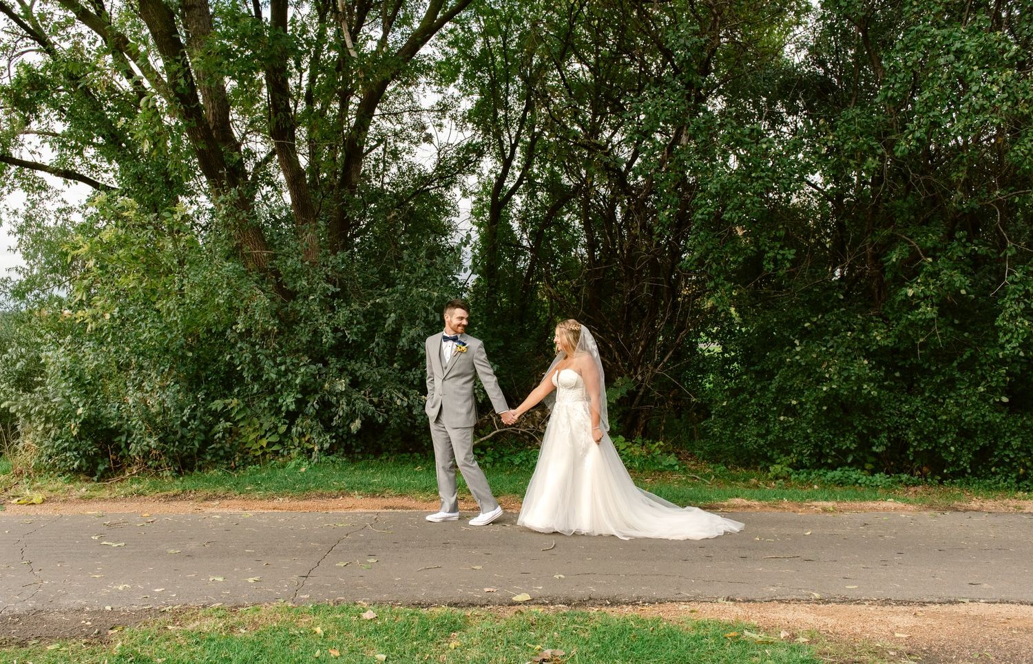 Central MN Wedding Photos | Bride and groom holding hands on a path surrounded by lush green trees on wedding day.