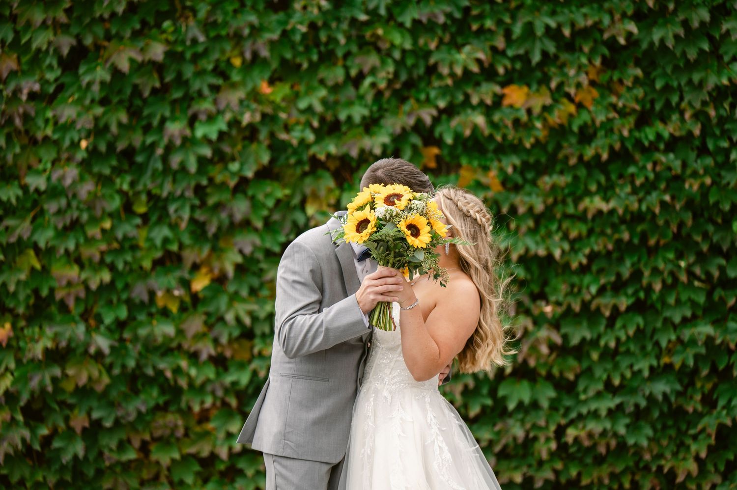 Central MN Wedding Photos | Bride and groom kiss behind vibrant sunflower bouquet against lush green ivy wall.