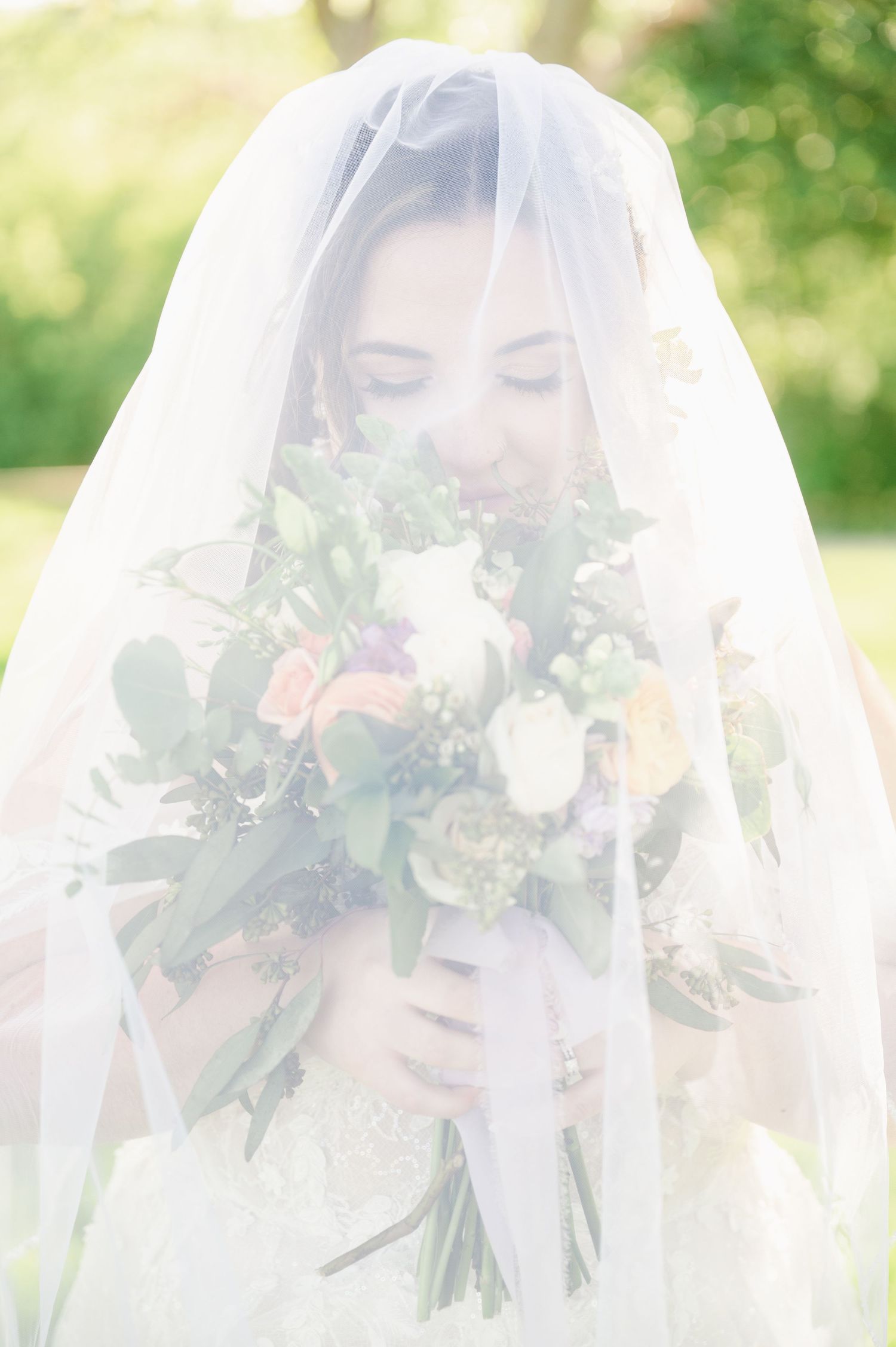 Central MN Wedding Photos - Bride in lace gown smelling colorful wedding bouquet under flowing white veil outdoors in garden setting.