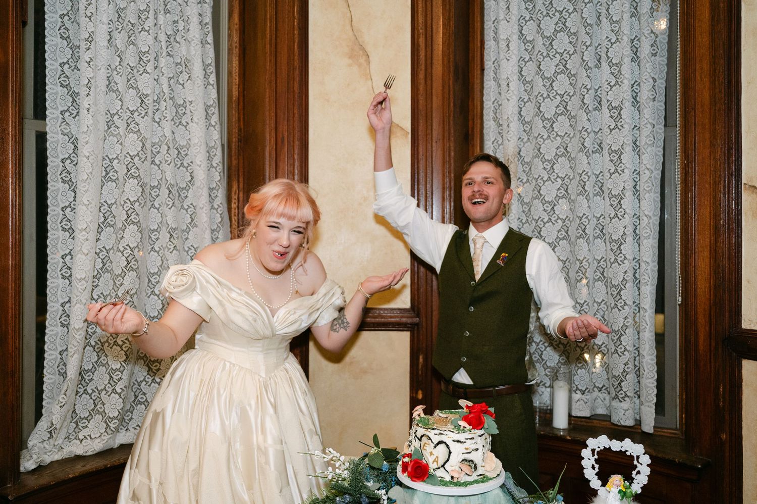 Central MN Wedding Photos | Bride and groom celebrate joyfully beside decorated wedding cake at indoor reception.