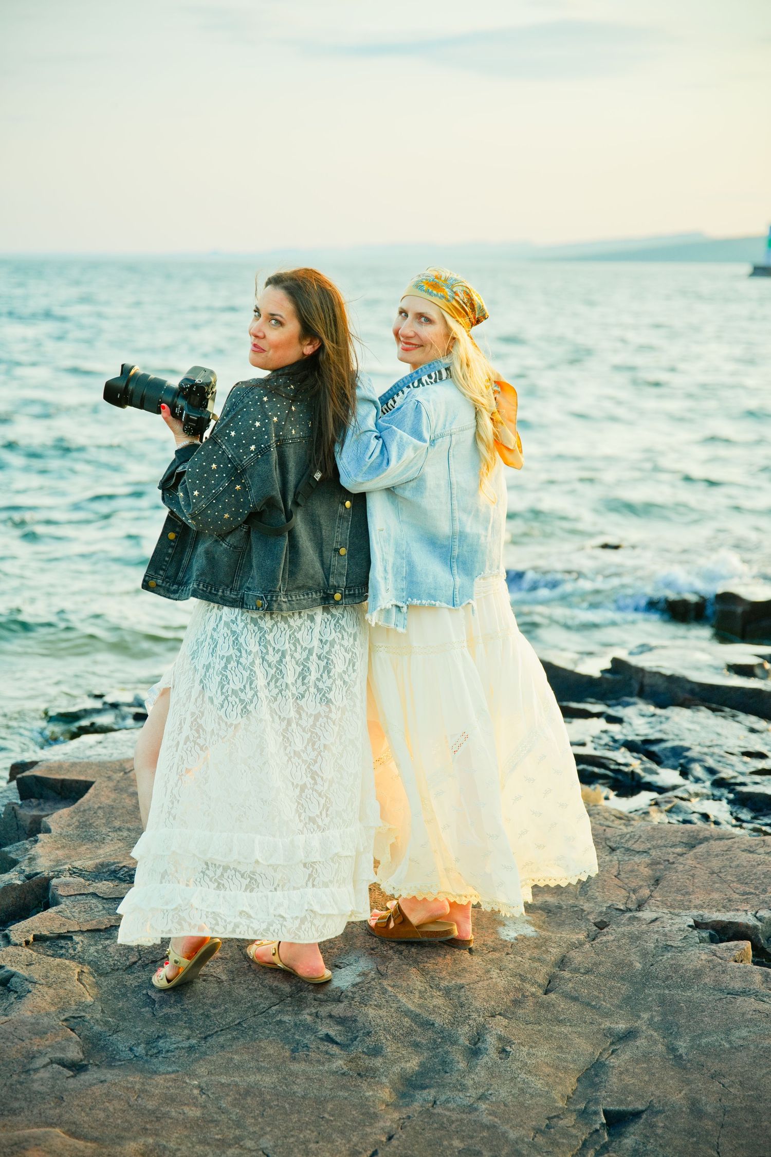 Two women in denim jackets and white lace skirts pose on rocky shoreline looking back, one holding a camera, with the sea behind.