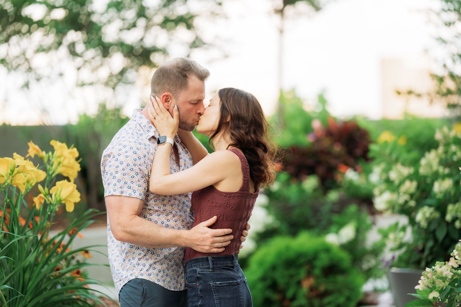 A couple shares a romantic moment in a lush garden setting surrounded by blooming flowers and greenery.