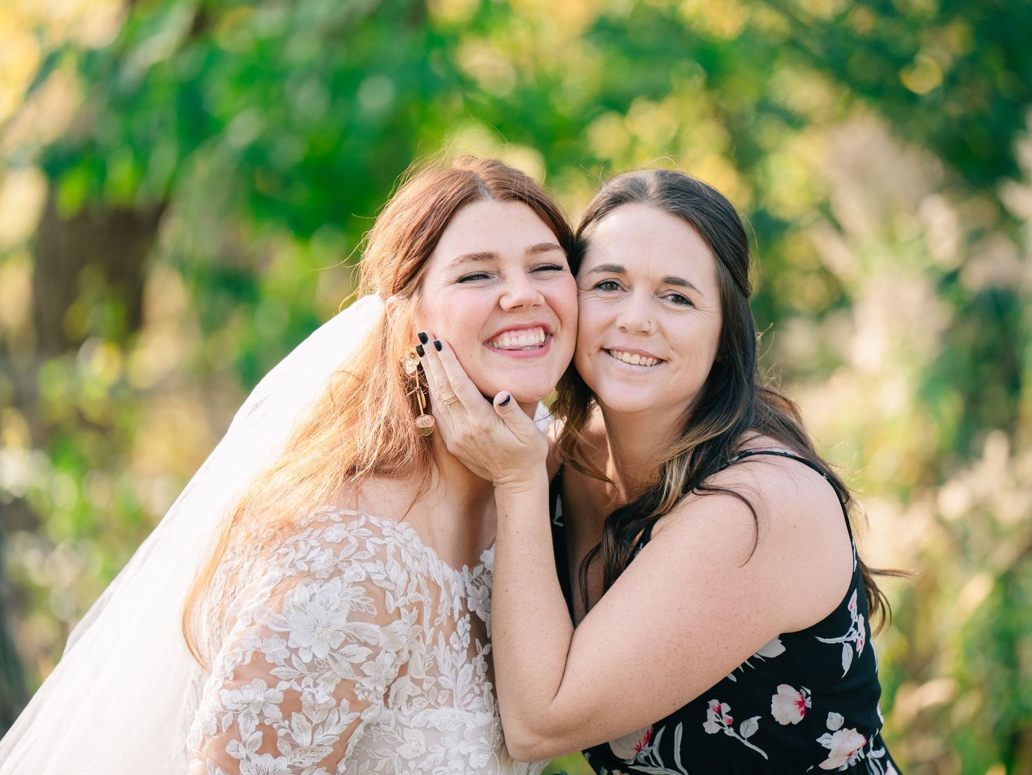 Smiling bride in lace veil embraces bridesmaid in black floral dress outdoors with green foliage background.
