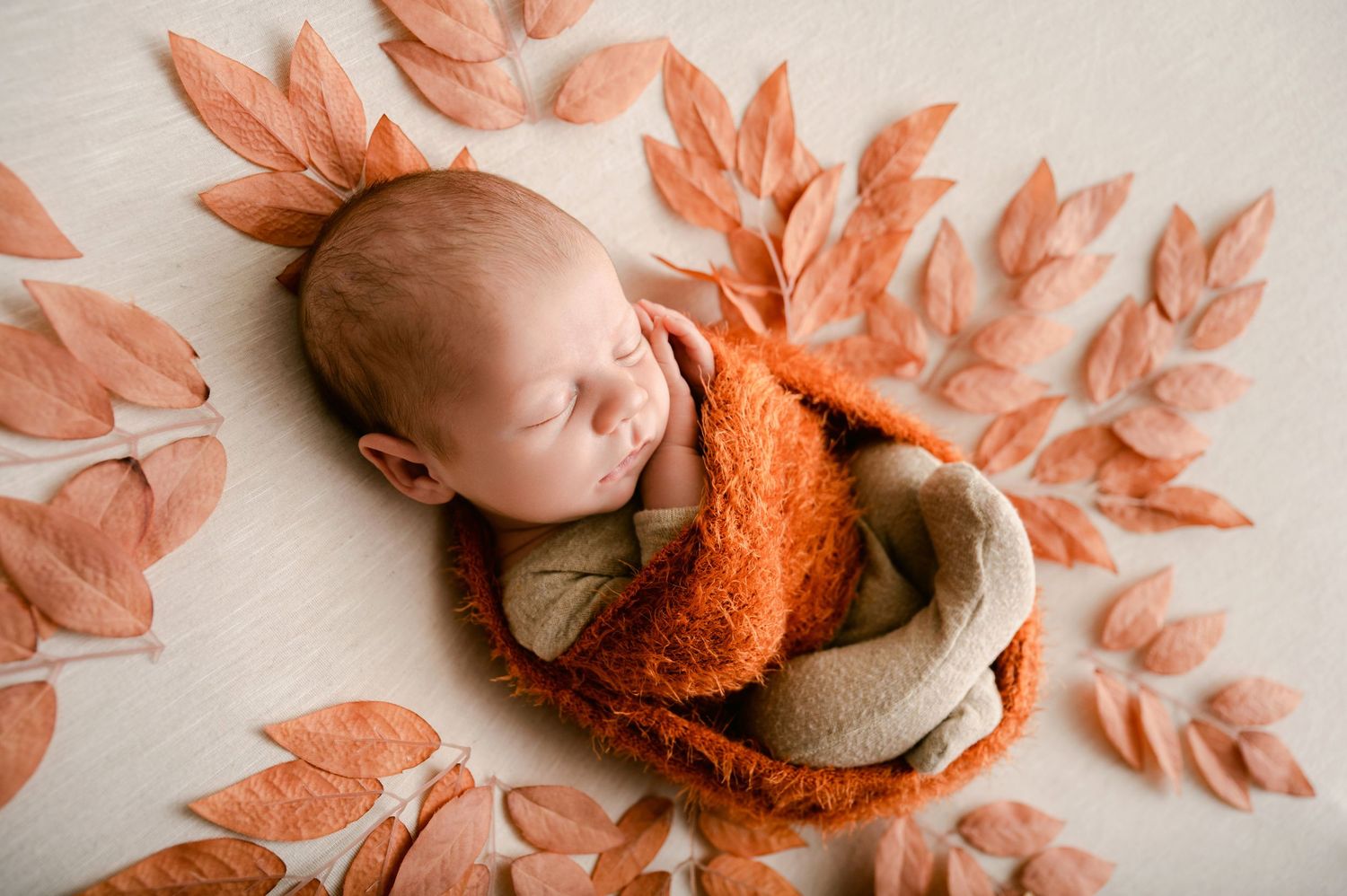 A sleeping baby adorned with autumn leaves lays nestled in a cozy orange blanket during a fall-themed photo shoot.