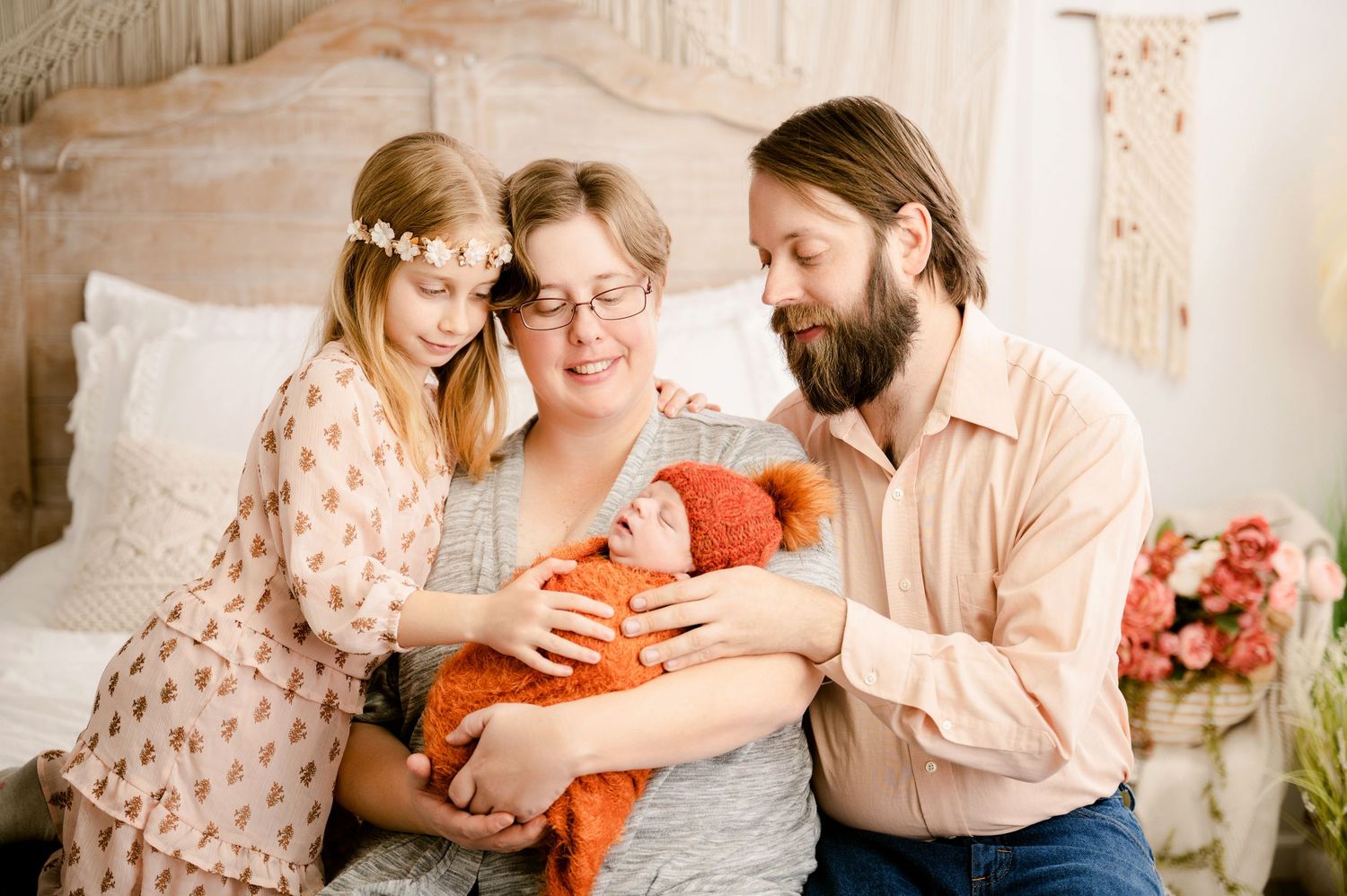 A family gathers together on a bed to admire their newborn baby wrapped in an orange blanket.