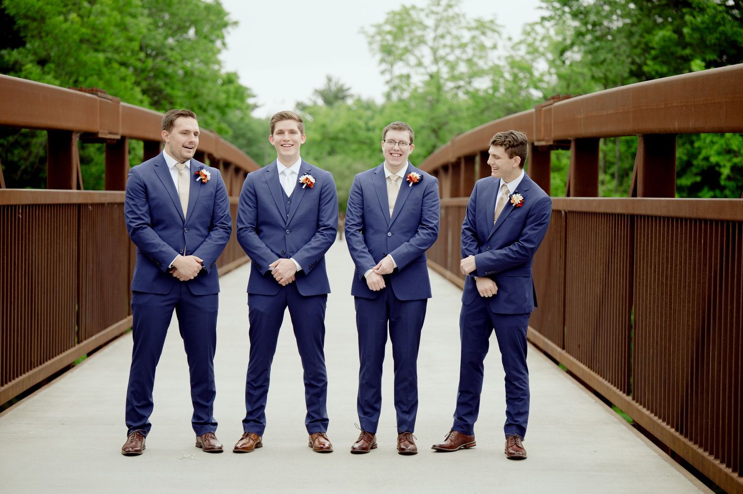 Four groomsmen in matching navy suits stand together on a wooden bridge surrounded by green foliage.