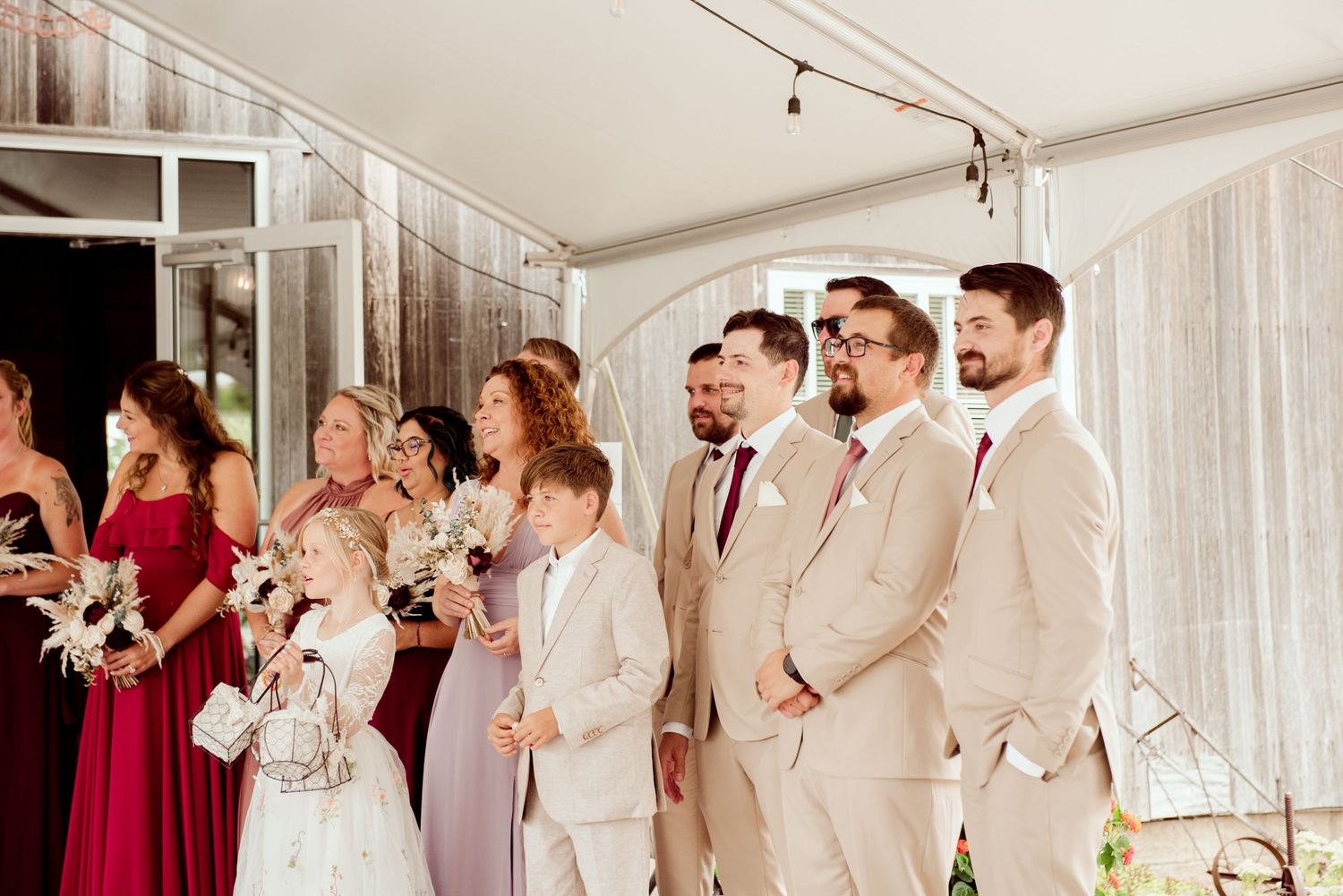 Wedding party lined up under a white tent with bridesmaids in red and groomsmen in beige suits.