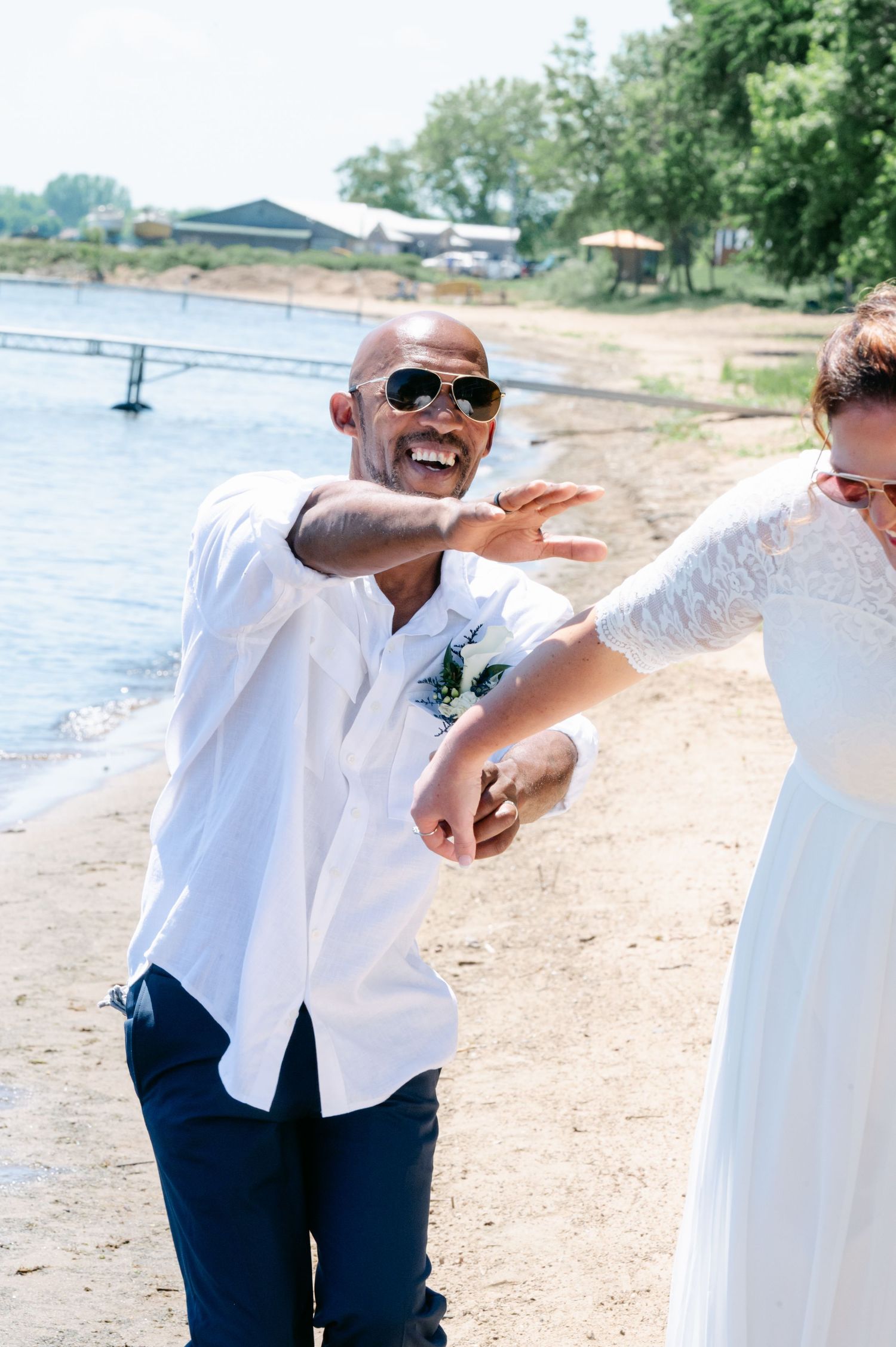Laughing groom in white shirt and sunglasses dancing with bride on sunny beach.