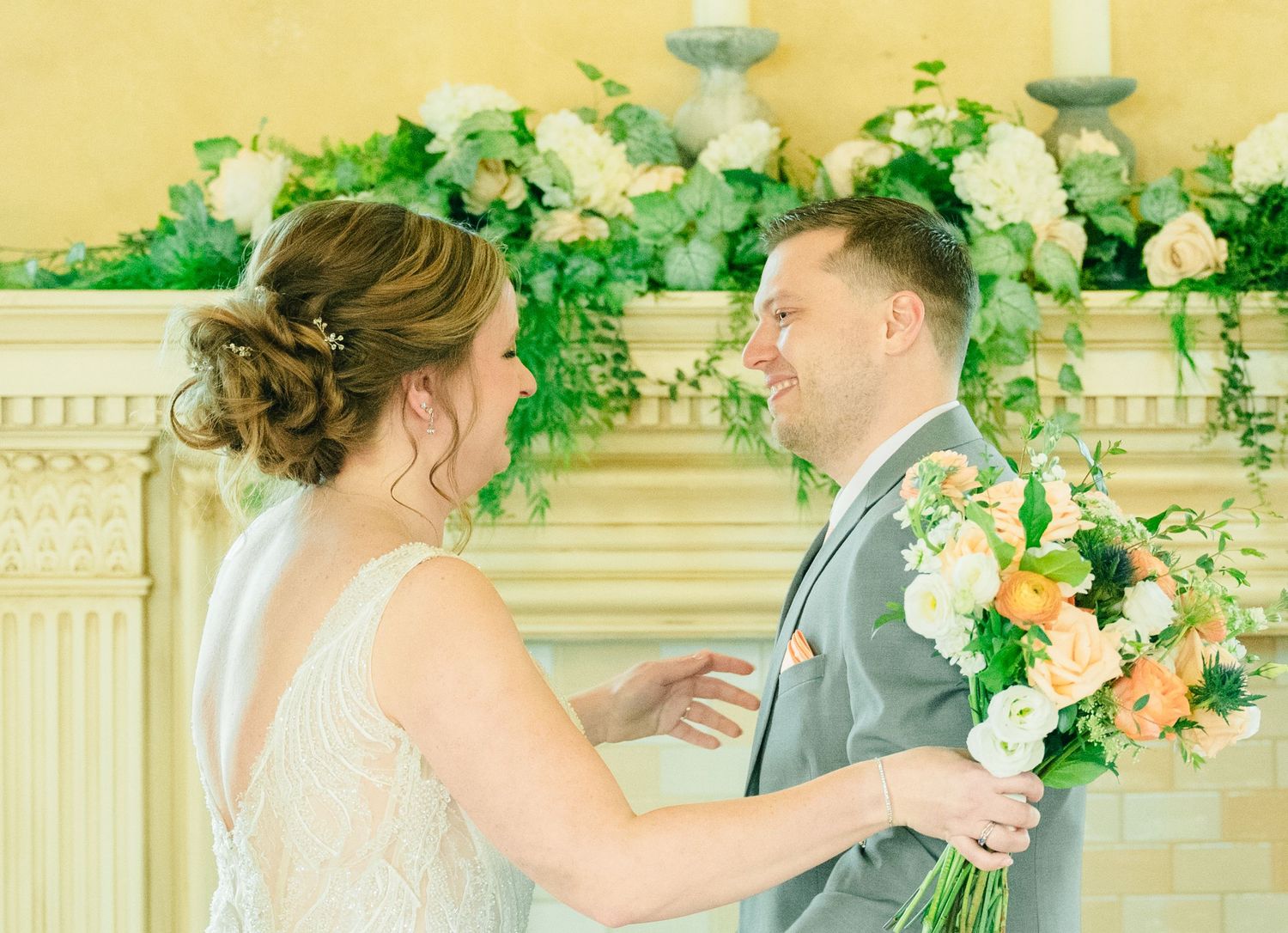 Bride and groom share a tender moment at altar decorated with lush greenery and peach floral arrangements.