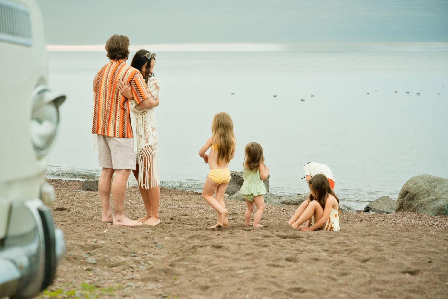 Family with three young children enjoying a peaceful beach day near a vintage van by the water.