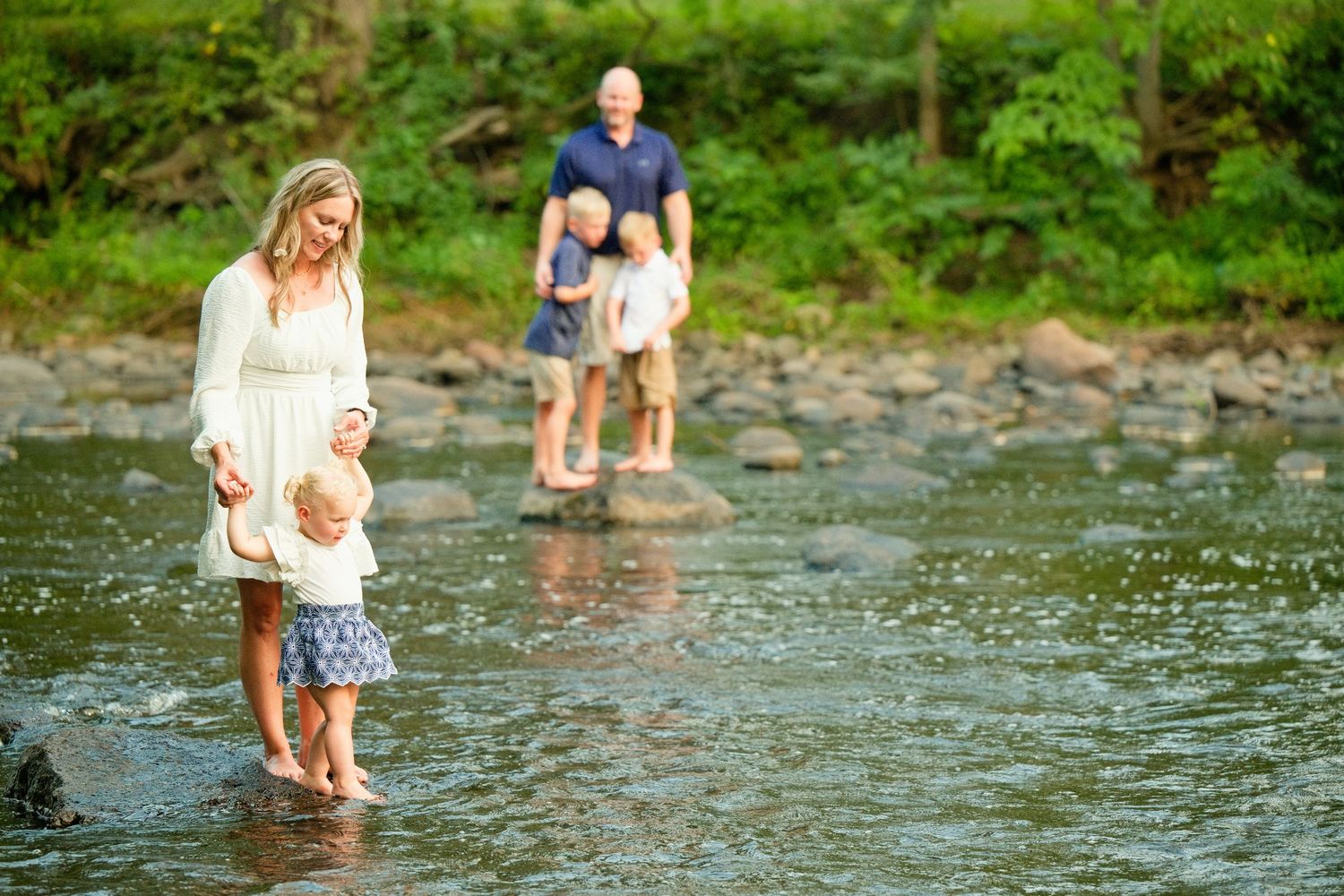 Family wading in a shallow creek, mother holding toddler's hand in foreground, couple with child on rocks behind.