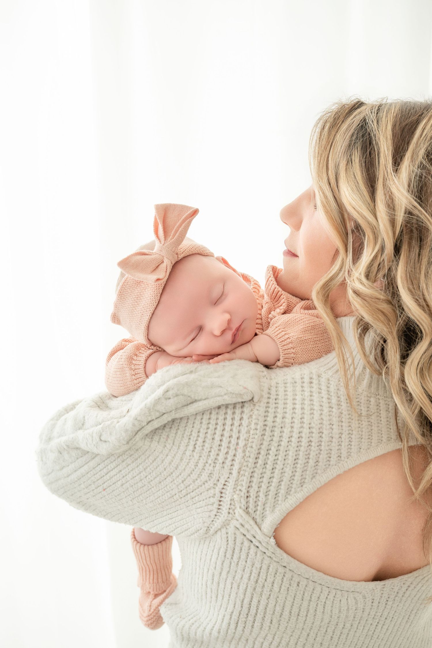 Blonde mother in gray sweater tenderly holds sleeping newborn baby girl wearing pink bow headband and outfit.