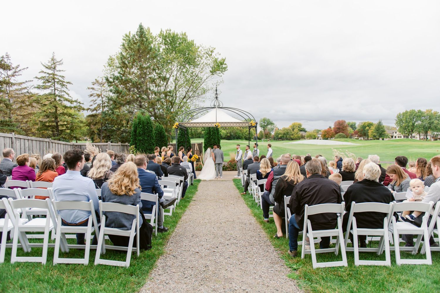 Central MN Wedding Photos - Outdoor wedding ceremony with white chairs on green lawn leading to elegant gazebo altar.