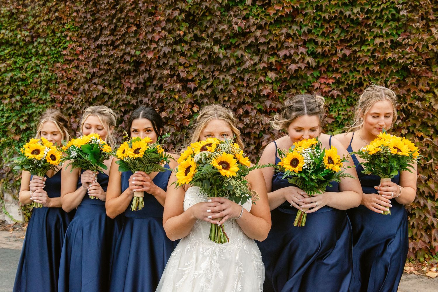 Central MN Wedding Photos | Bridesmaids in navy dresses hold sunflower bouquets in front of ivy-covered wall.
