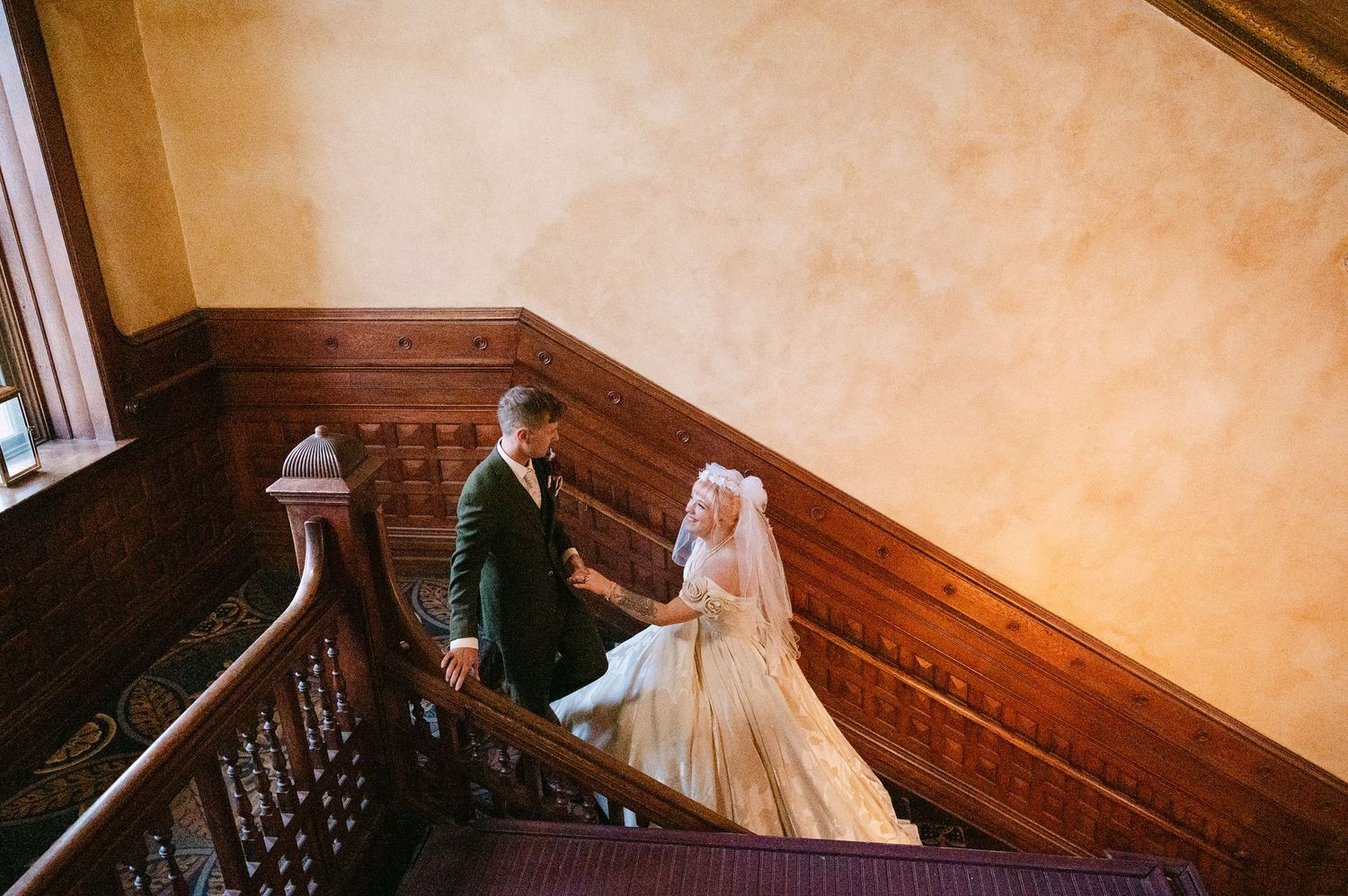 Central MN Wedding Photos | Bride and groom hold hands walking up grand wooden staircase in elegant historic venue.