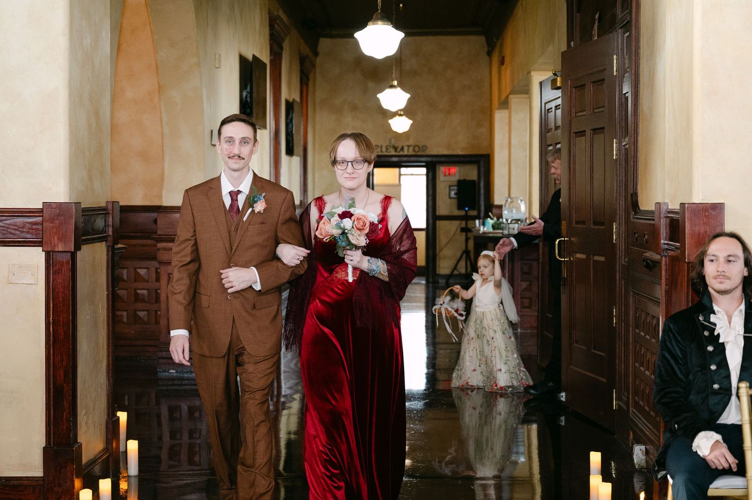 Central MN Wedding Photos | Wedding party in vintage hallway with groom in brown suit escorting woman in red velvet gown.
