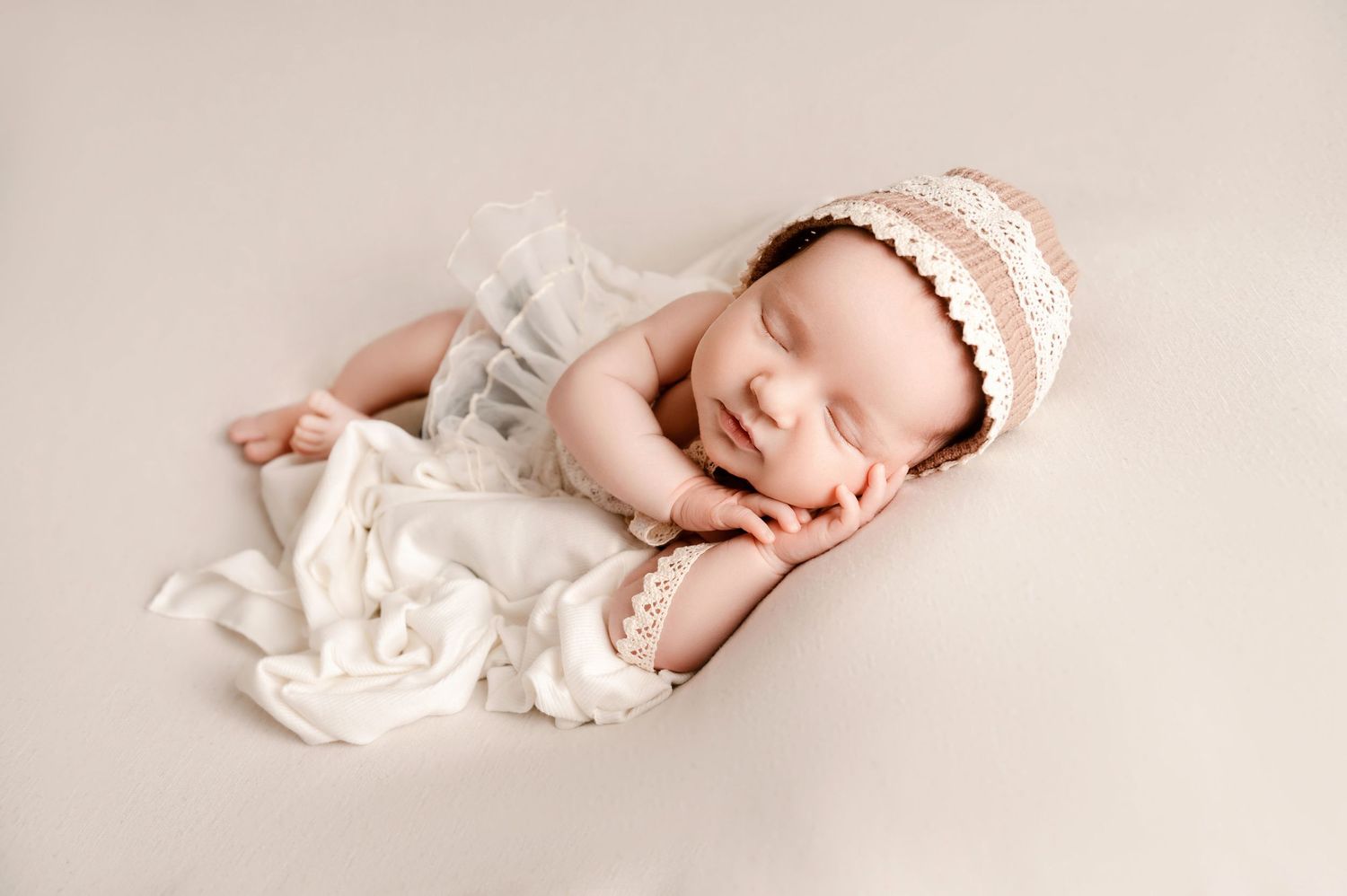 Peaceful newborn baby sleeping in white wrap and lace bonnet on beige backdrop.