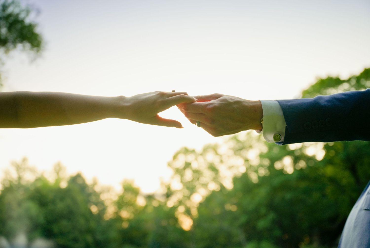 Two people gently touching hands outdoors with green trees and soft sunlight in the background.