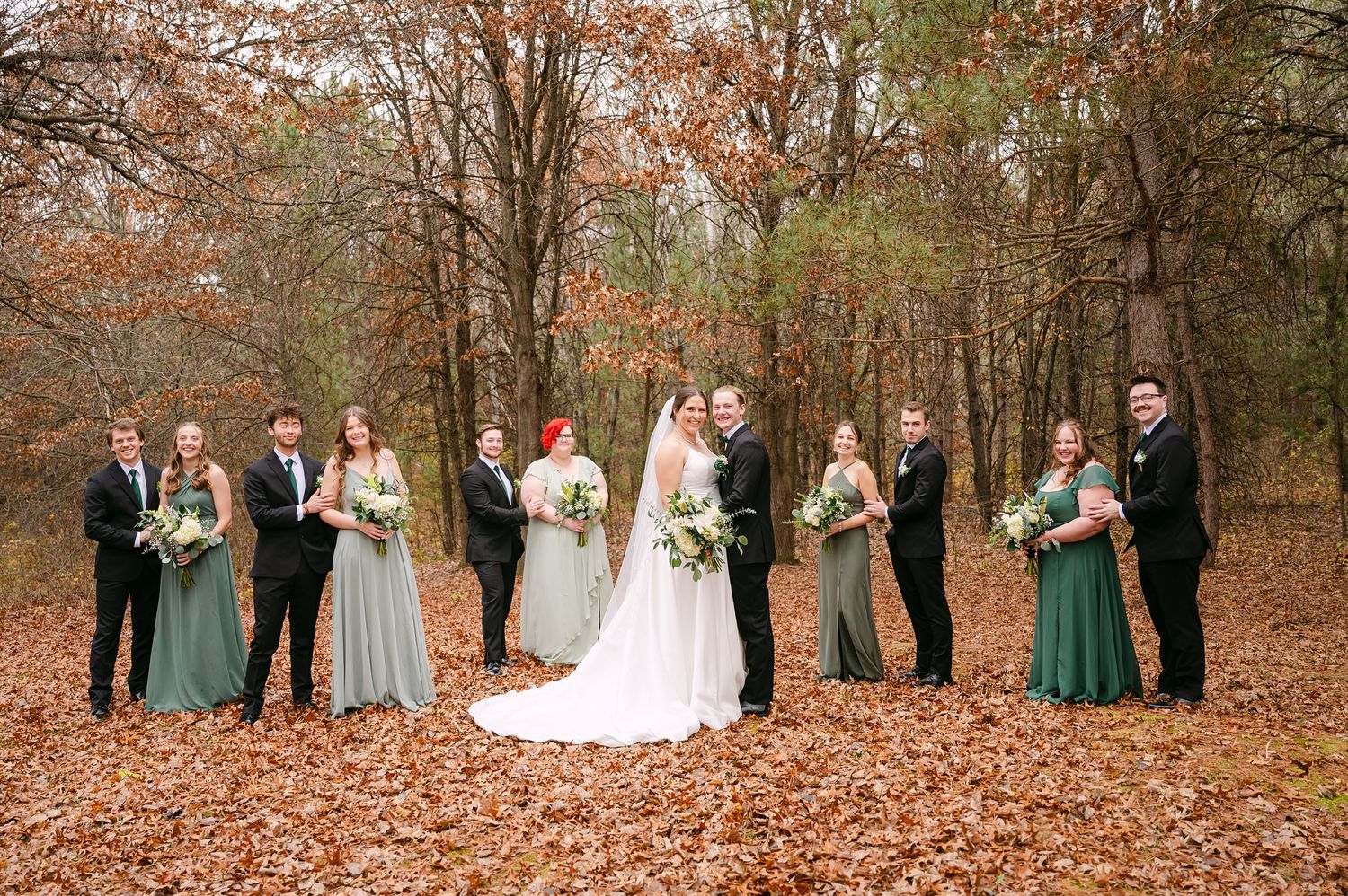 In central Minnesota a Wedding party poses for autumn photos in a forest setting with fallen leaves and sage green bridesmaid dresses.