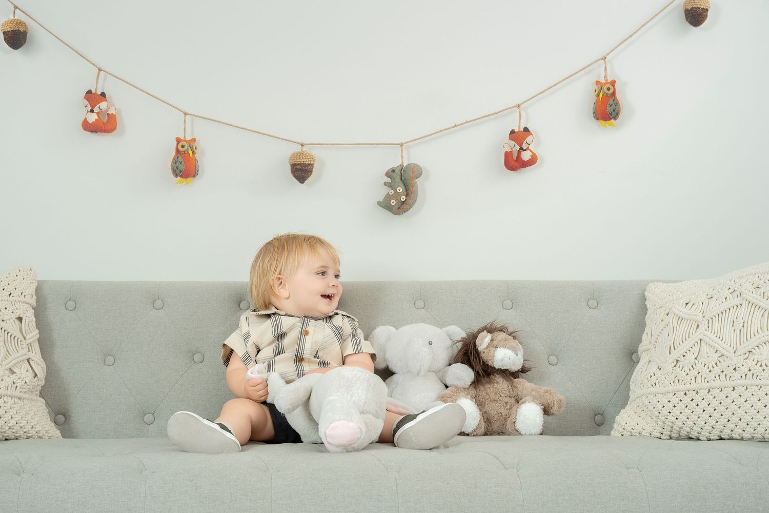Central Minnesota Toddler sitting on gray couch with stuffed animals under festive fall garland decoration. Minnesota milestone photographer