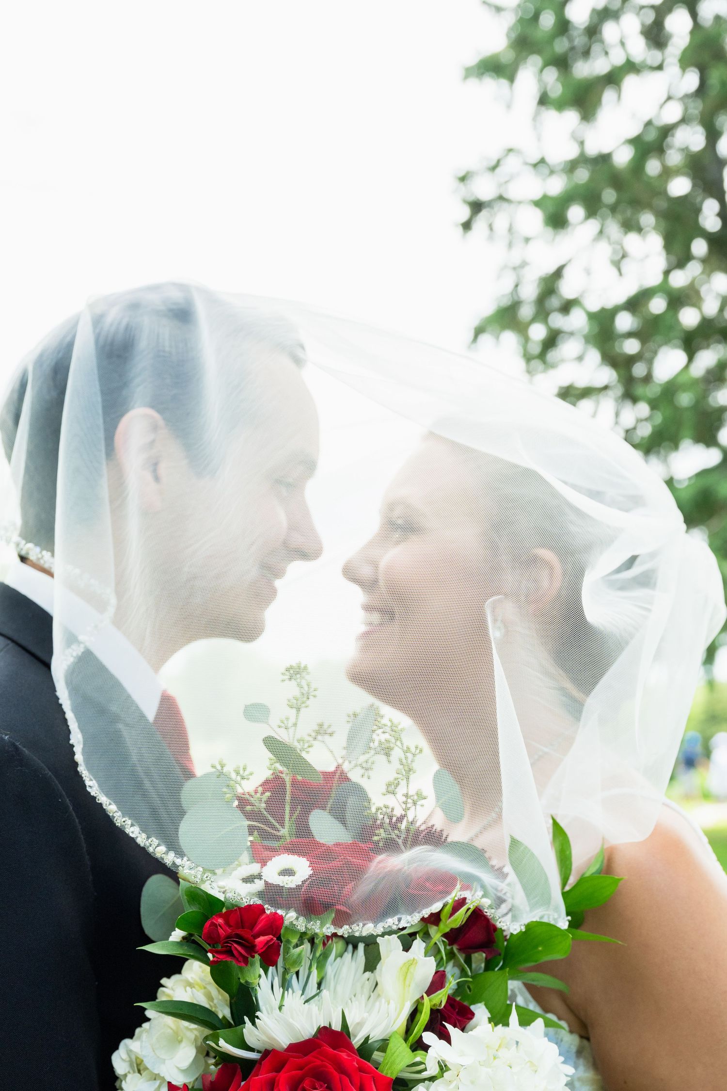 Bride and groom share a romantic moment under a sheer veil, smiling at each other while holding a red bouquet.