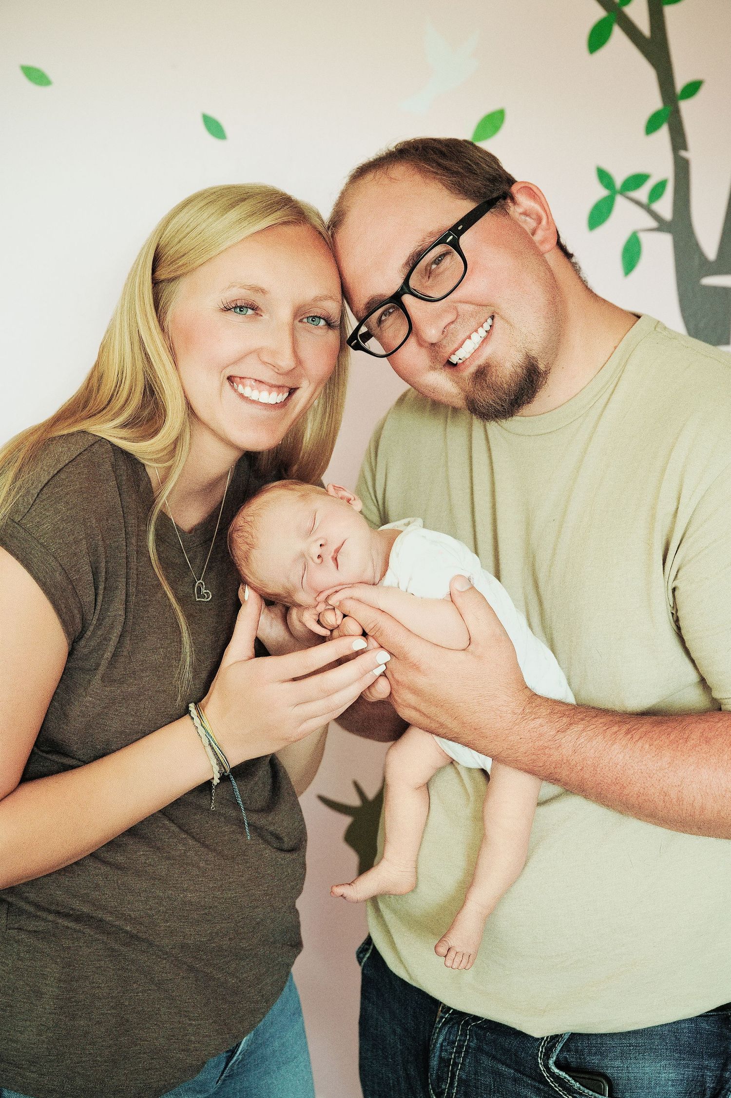 Smiling young couple holding their sleeping newborn baby in a nursery with tree wall decal.