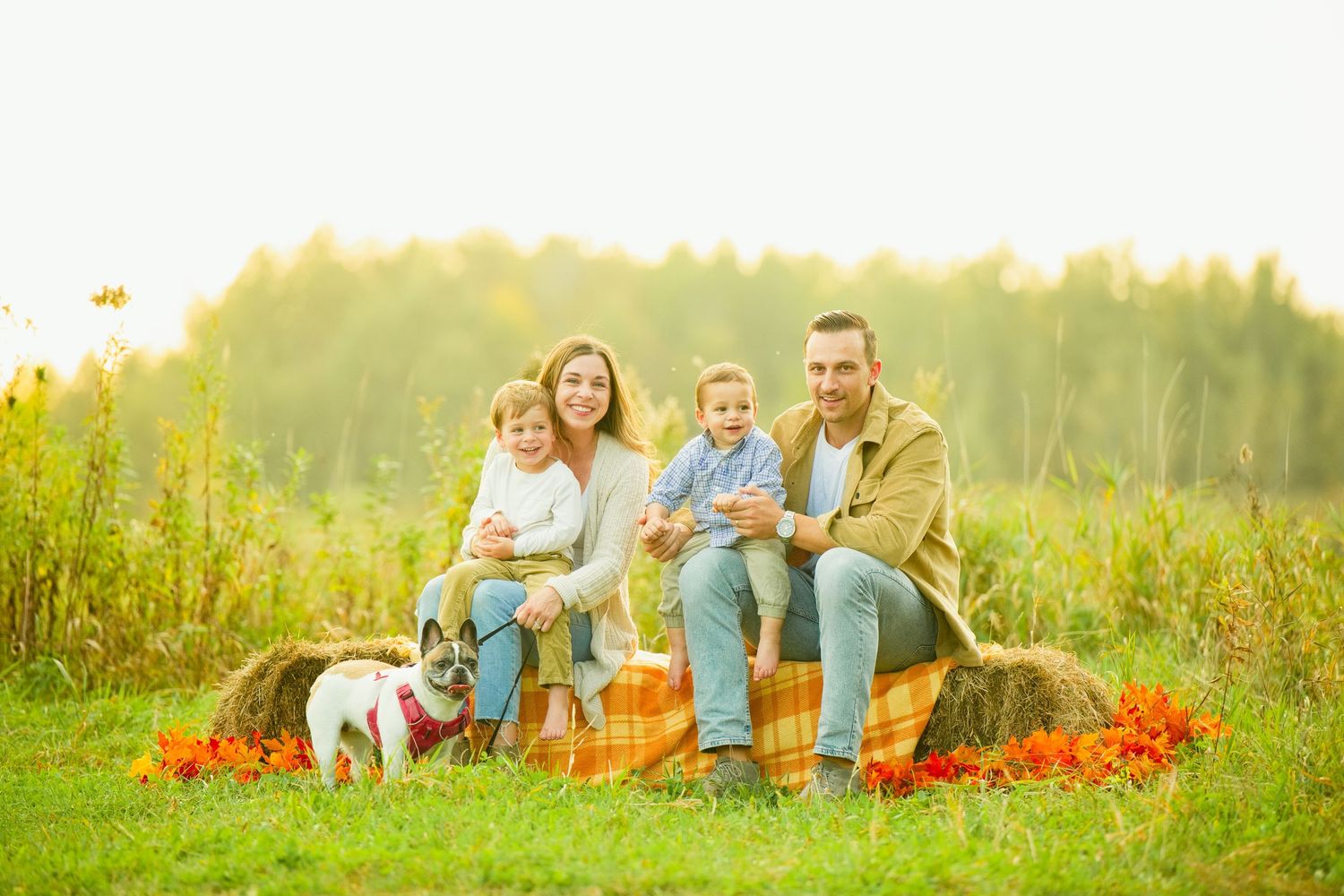 Happy family with two toddlers and a dog sitting on hay bales in a sunny autumn field.