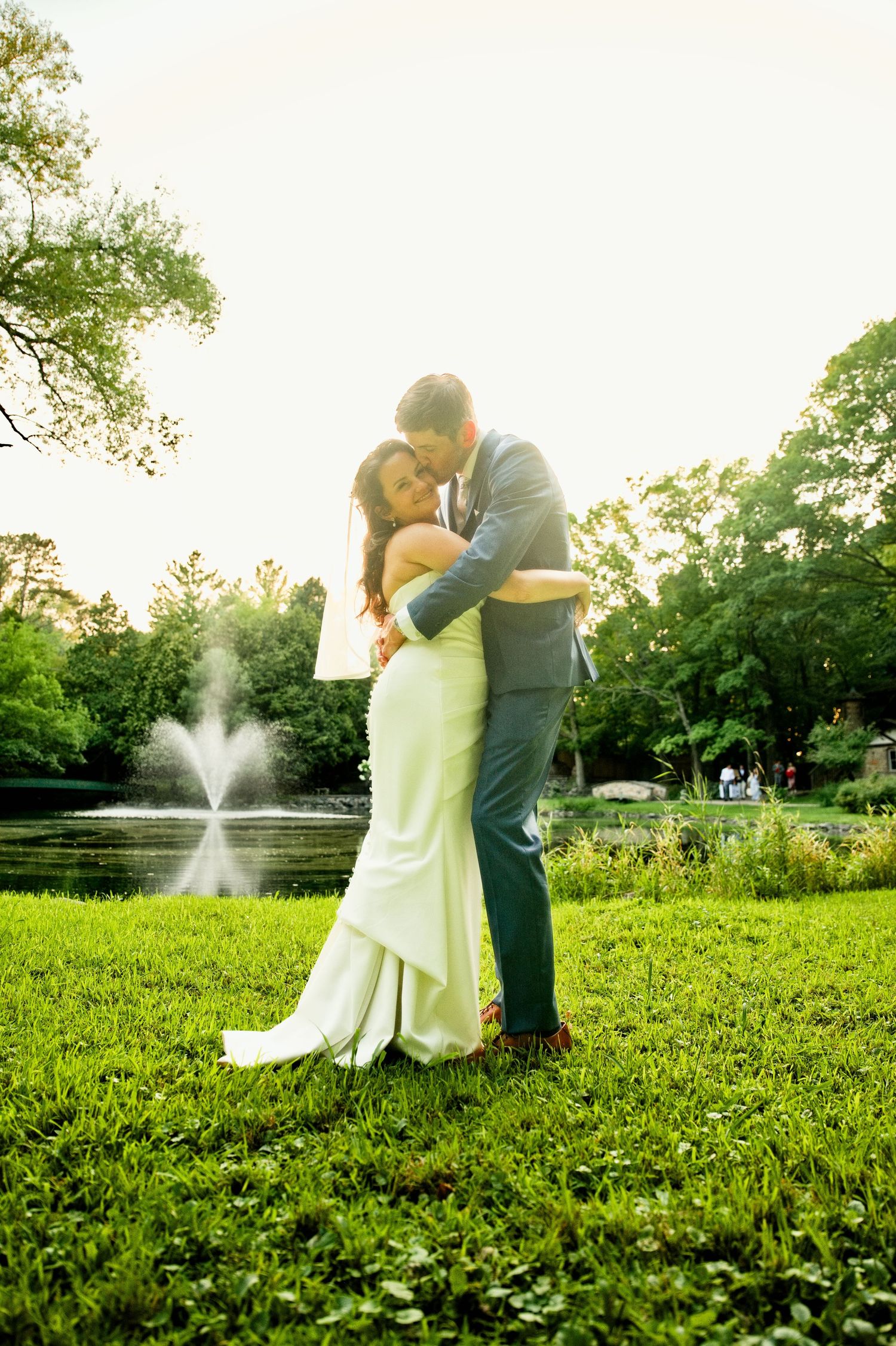 Bride and groom embrace on lush green grass near a fountain at golden hour in an outdoor wedding portrait.